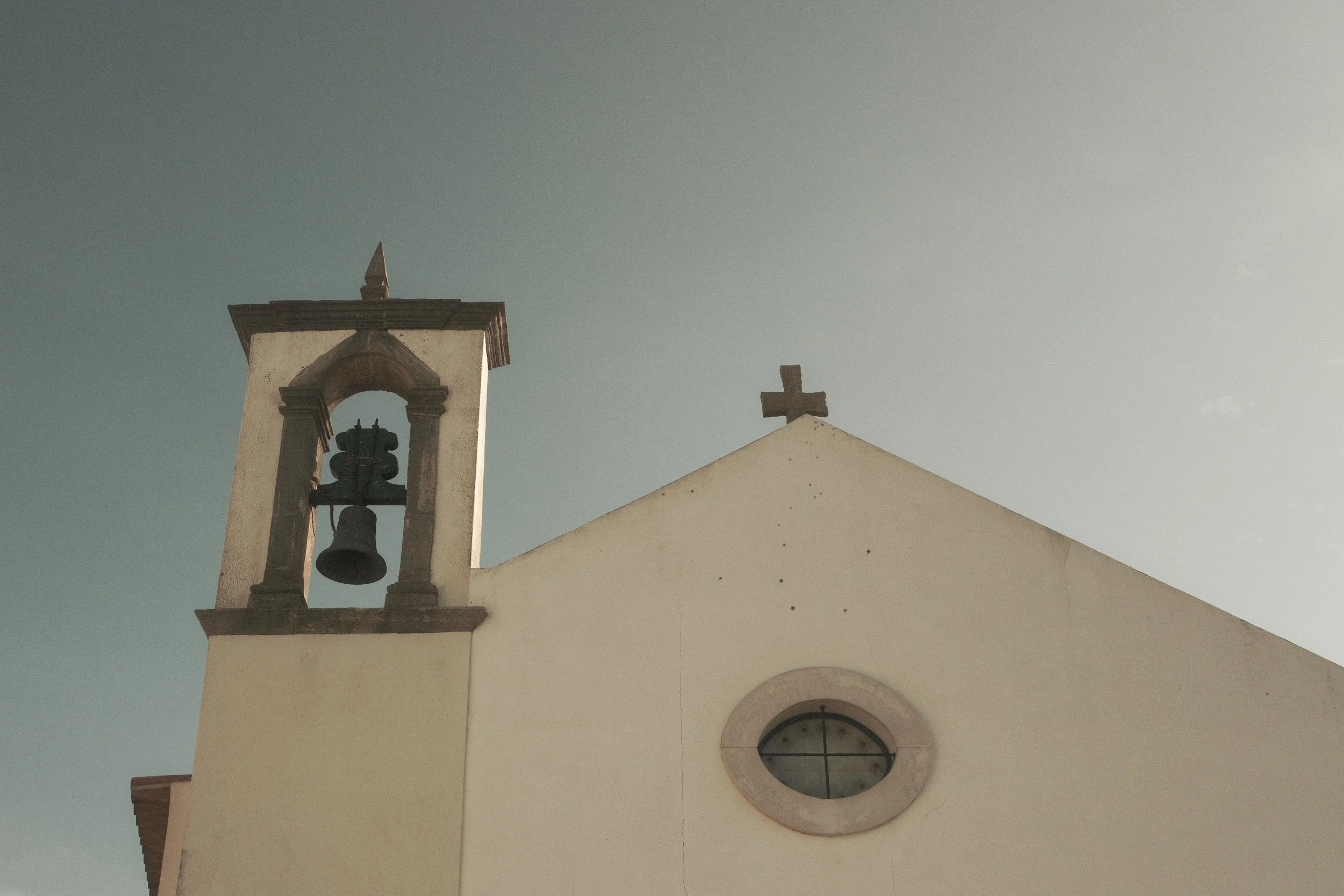 A church with a small bell tower on the left, a simple cross on top of the roof, and a round window below it, set against a pale sky.