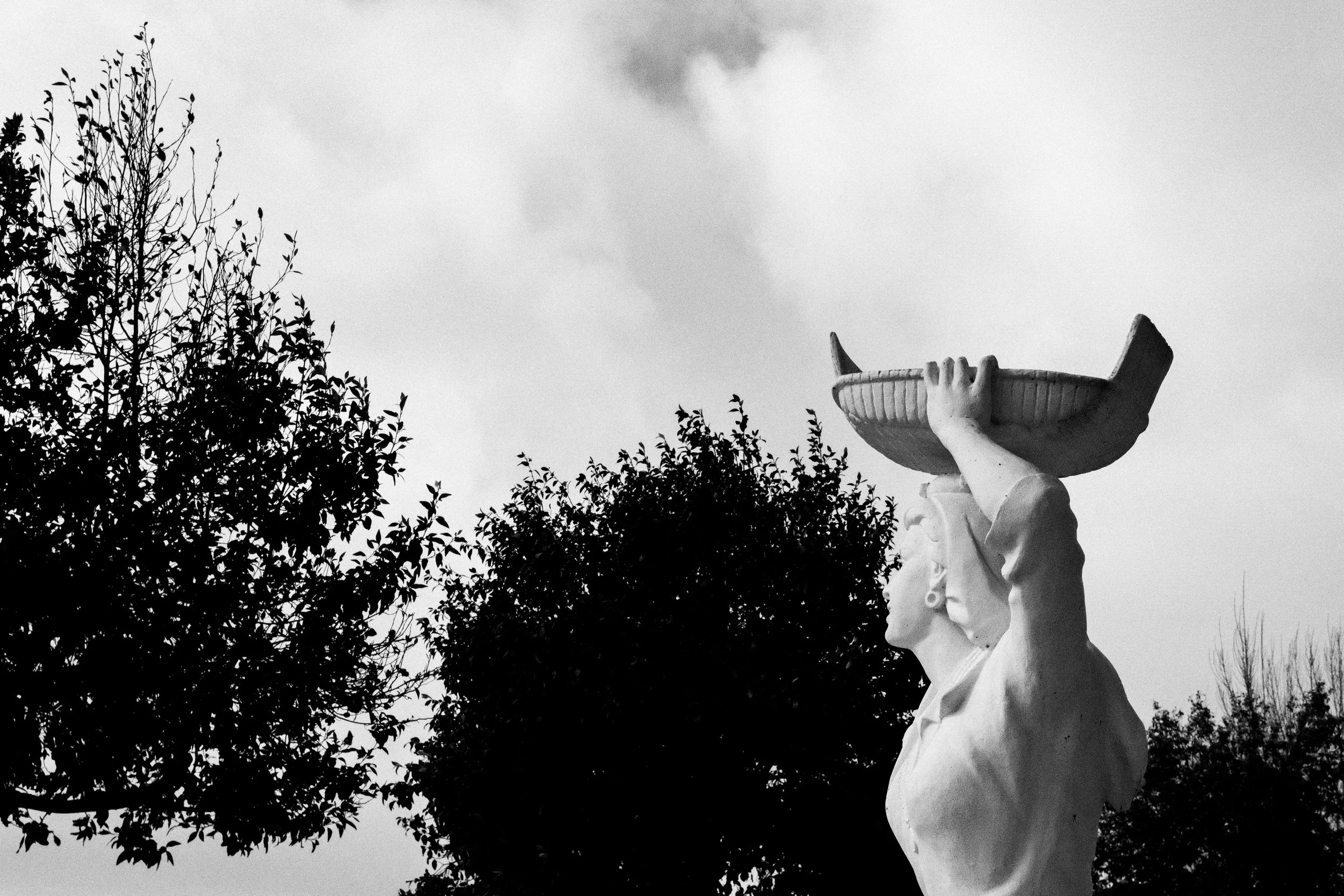 A statue of a woman holding a large shallow bowl above her head, with trees and a cloudy sky in the background.