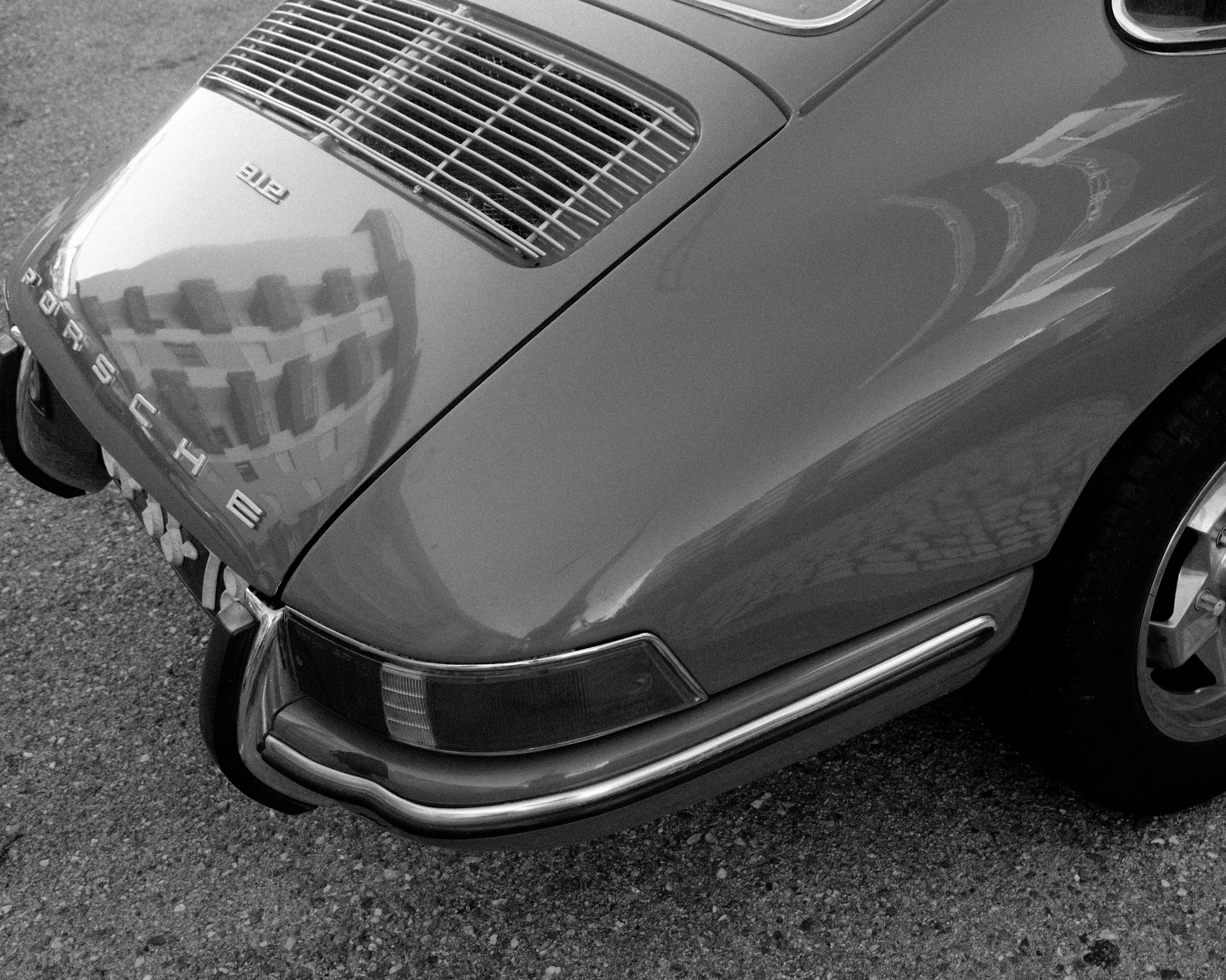 Close-up of the front left of a vintage Porsche 912 sports car, black and white photo, showing the hood, headlight, and part of the wheel, with reflections of buildings on the car's surface.