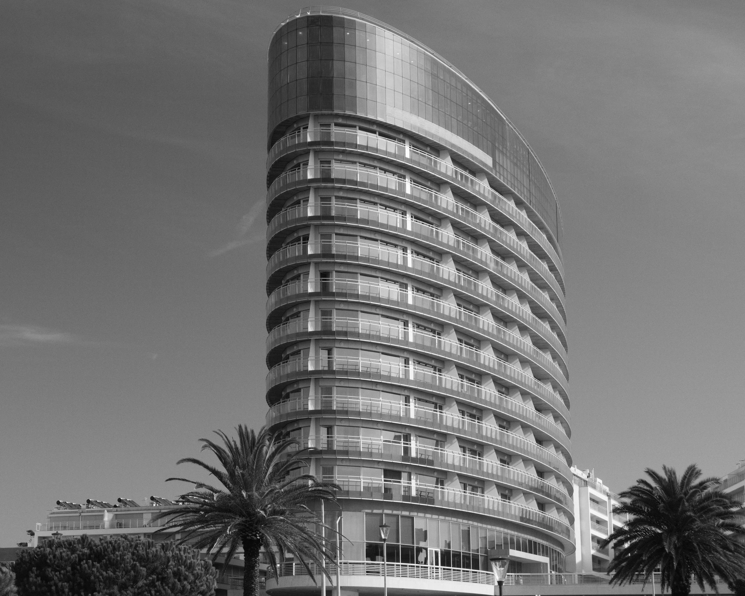 A modern, high-rise building with curved glass exterior and multiple balconies, surrounded by palm trees.