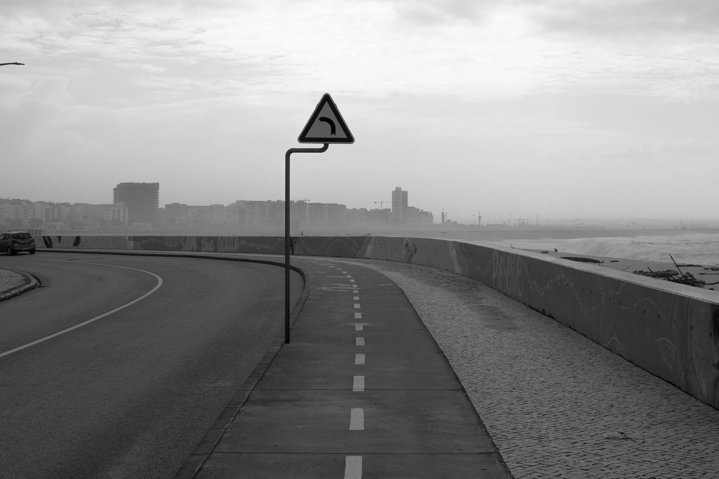 A black-and-white photograph of a coastal road with a sidewalk and a curved traffic sign indicating a left turn, overlooking an ocean and city skyline in the distance.