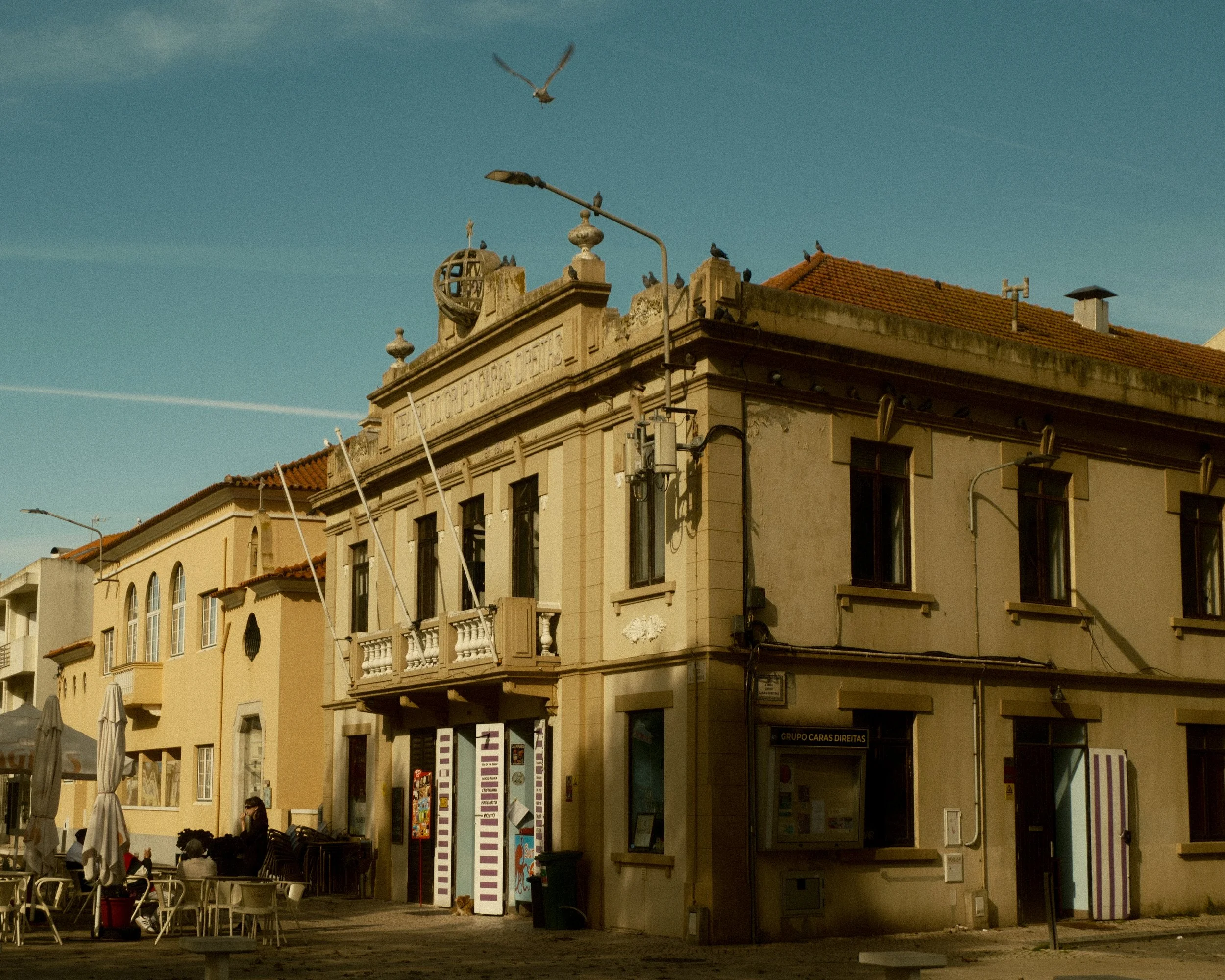 A corner building with classical architecture, featuring a decorative string of Latin text, a small balcony, and a globe sculpture on the roof. There are several windows, and a street scene in front with people sitting at tables under umbrellas, and 
