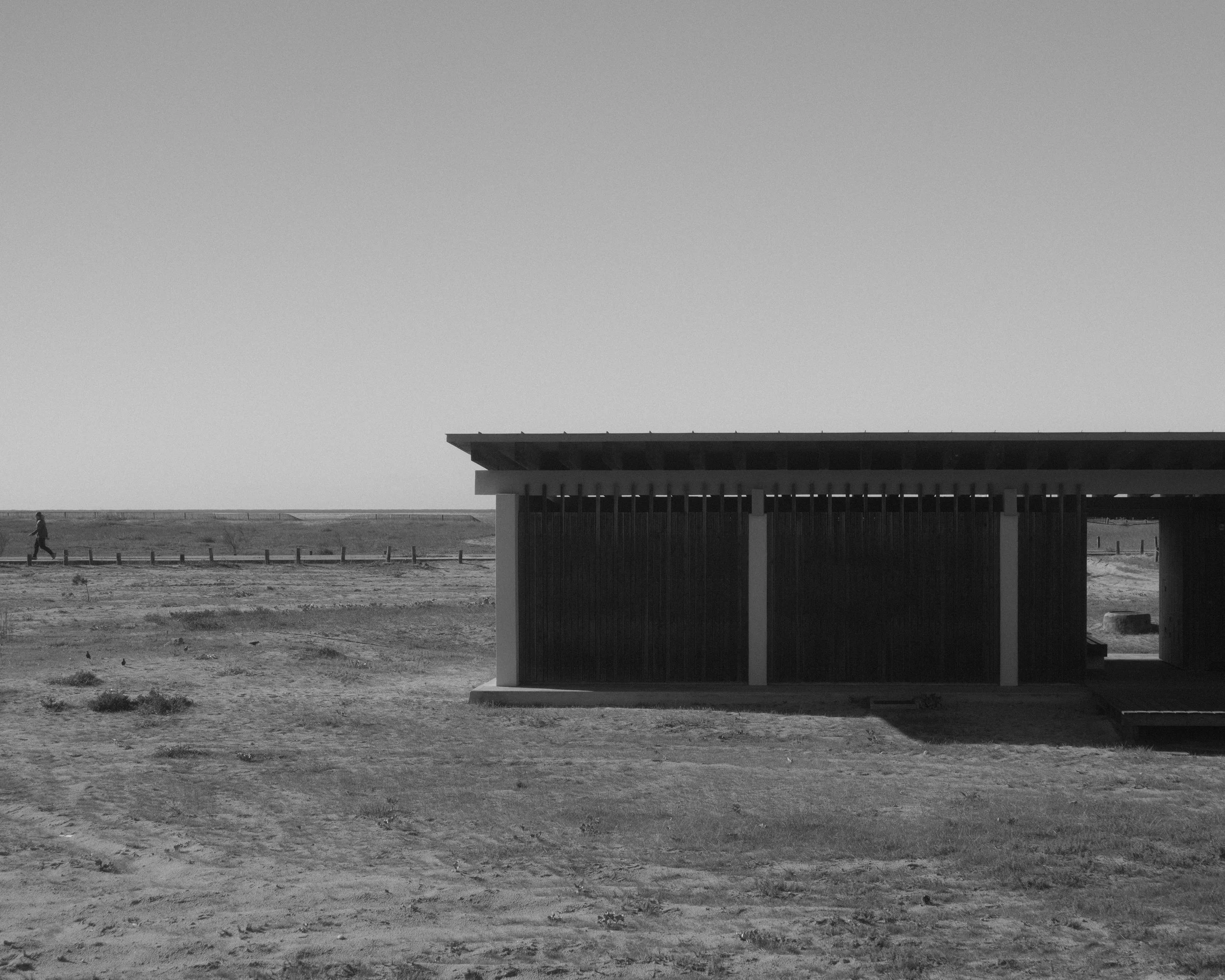 A black and white photo of a modern building with vertical slats and a sloped roof. A person is walking on a dirt path in the distance, under a large, clear, open sky.