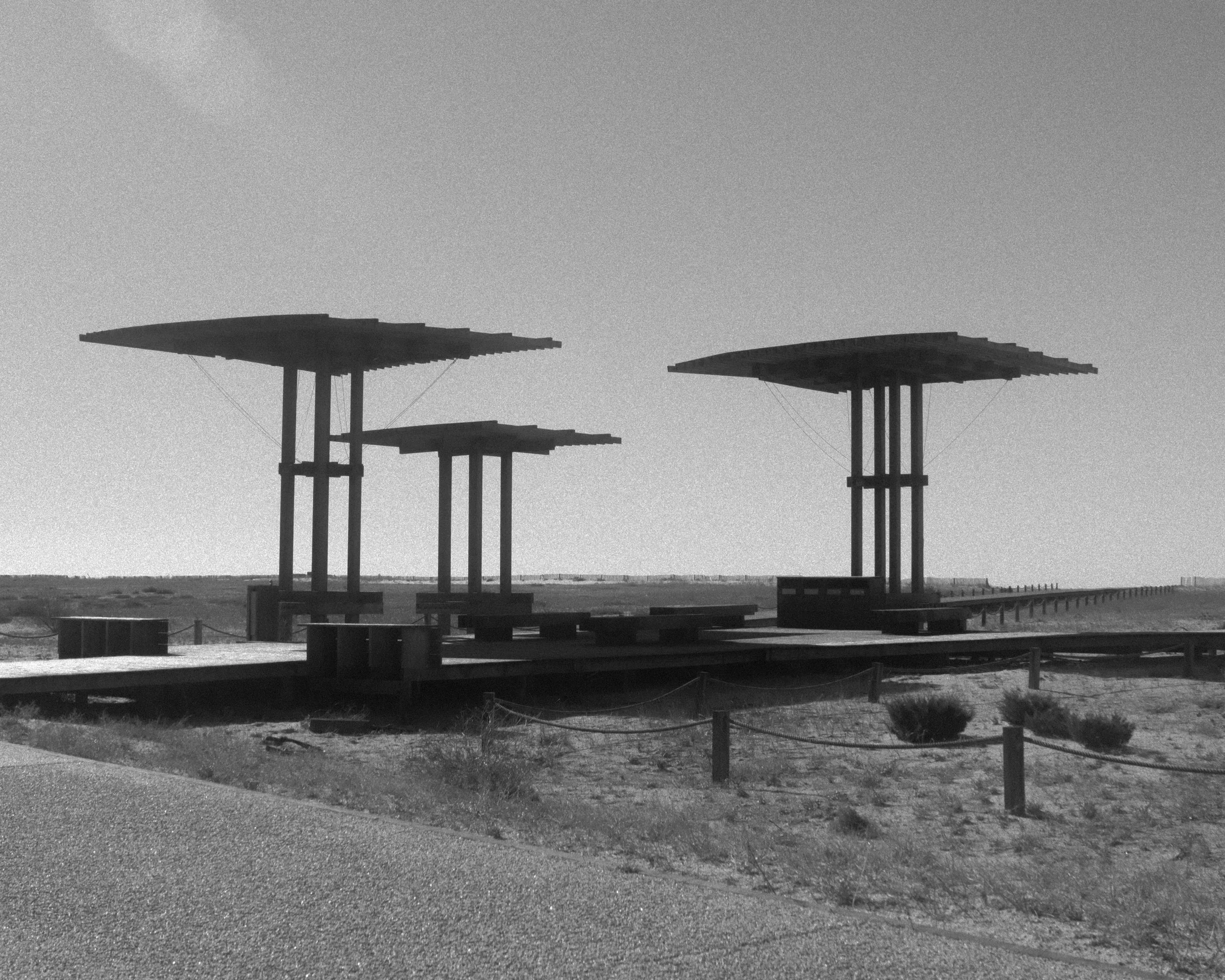 Black and white photo of a wooden promenade with large, modern, sculptural shade structures in a beach or desert setting.