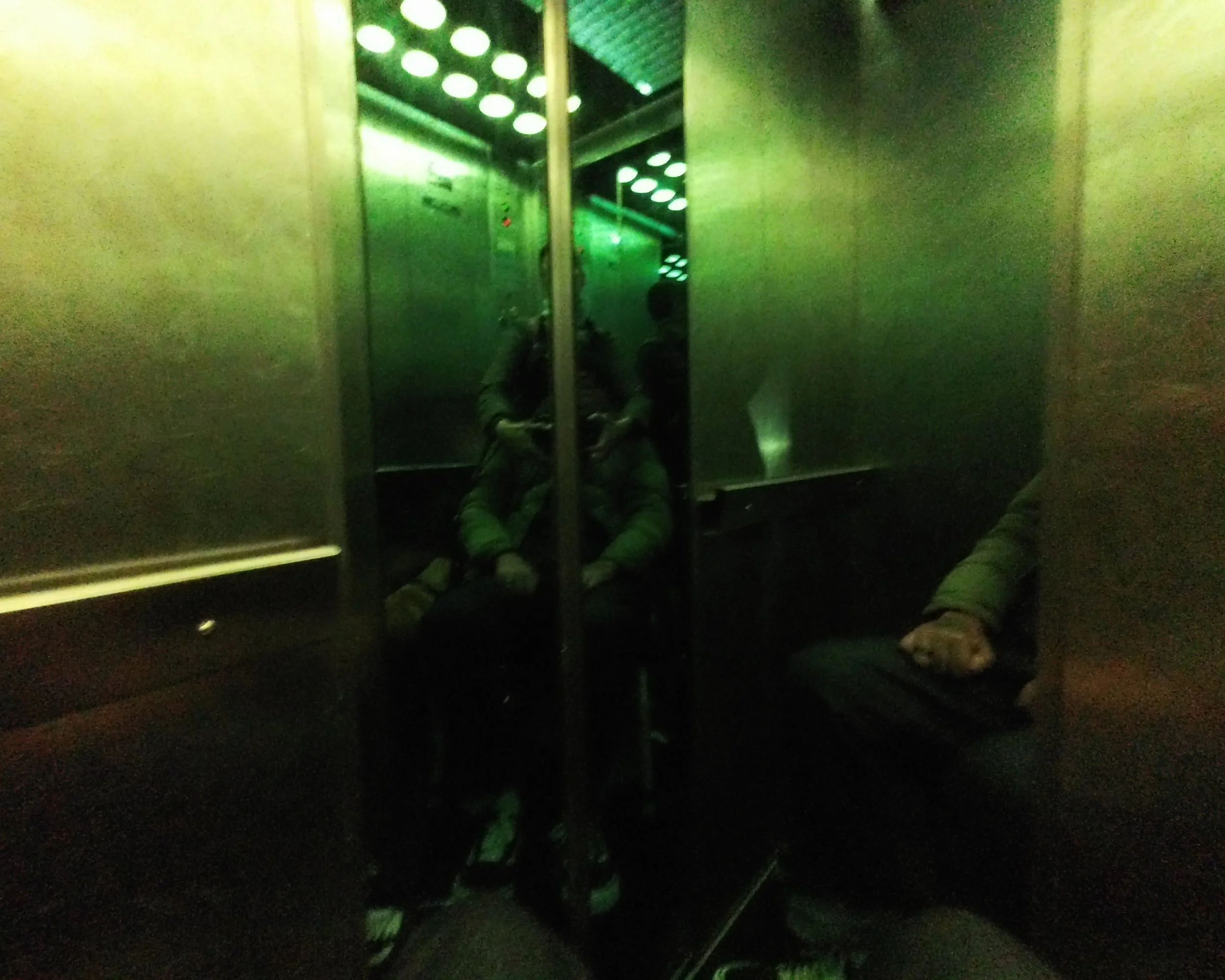 Two people sitting in an elevator with green lighting, their reflections visible on the metallic walls.