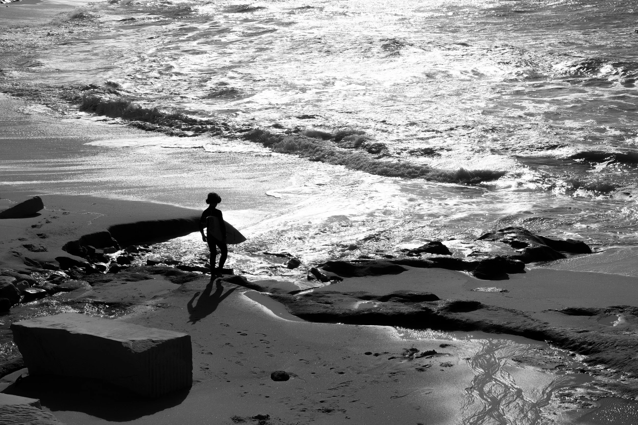 A silhouette of a person carrying a surfboard walks along a rocky beach with waves crashing in the background, seen in black and white.