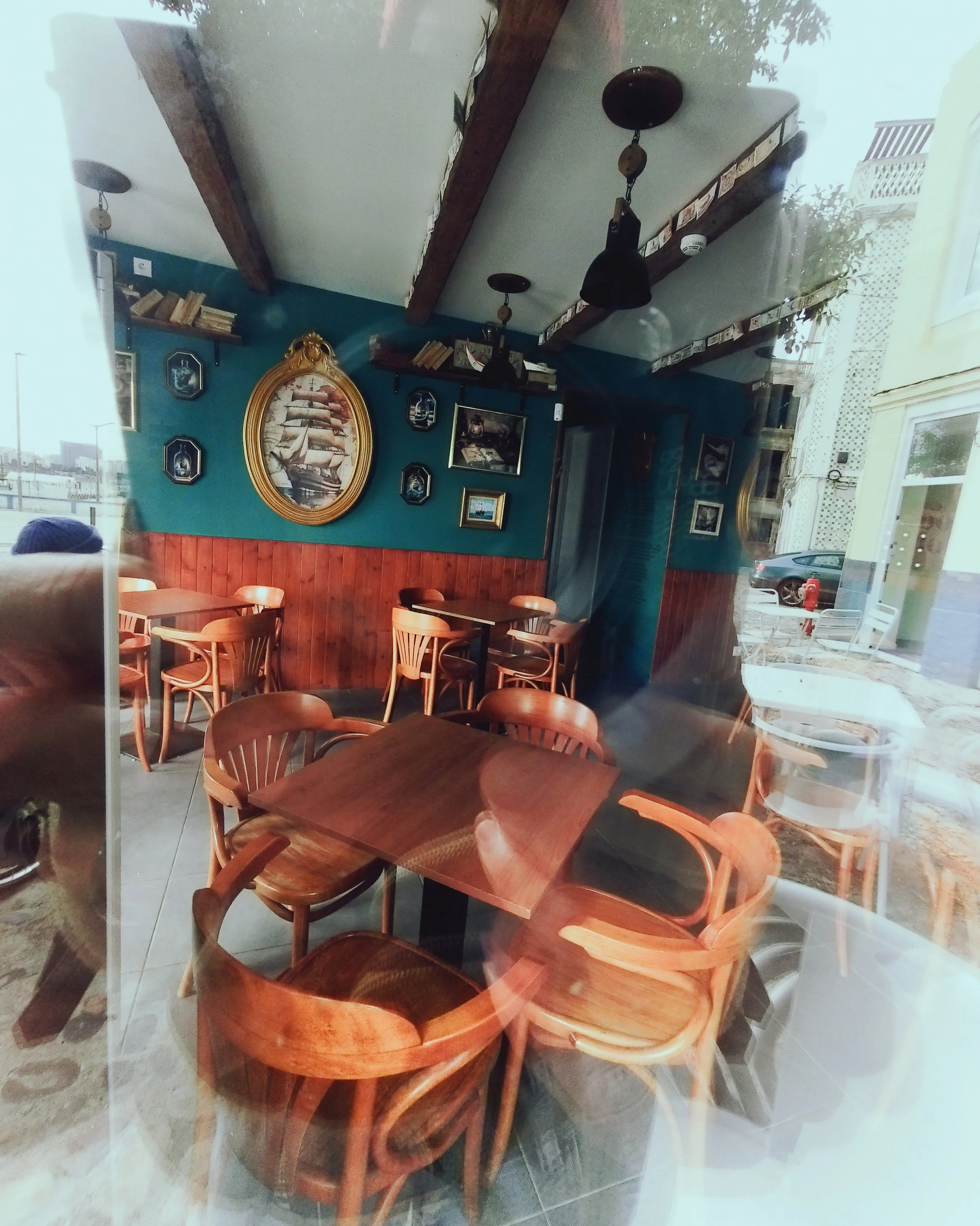 A cozy café interior with wooden tables and chairs, framed artwork and decorations on teal walls, and a ceiling with wooden beams.