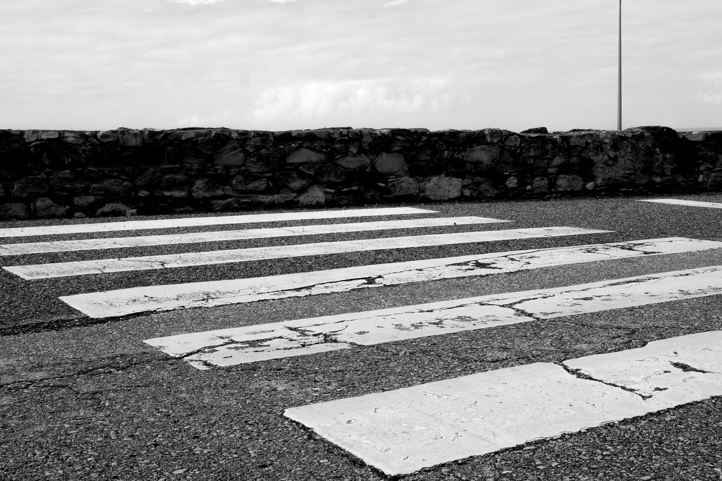 A worn pedestrian crosswalk with chipped white stripes on a rough asphalt road, a stone wall in the background, and cloudy sky overhead.