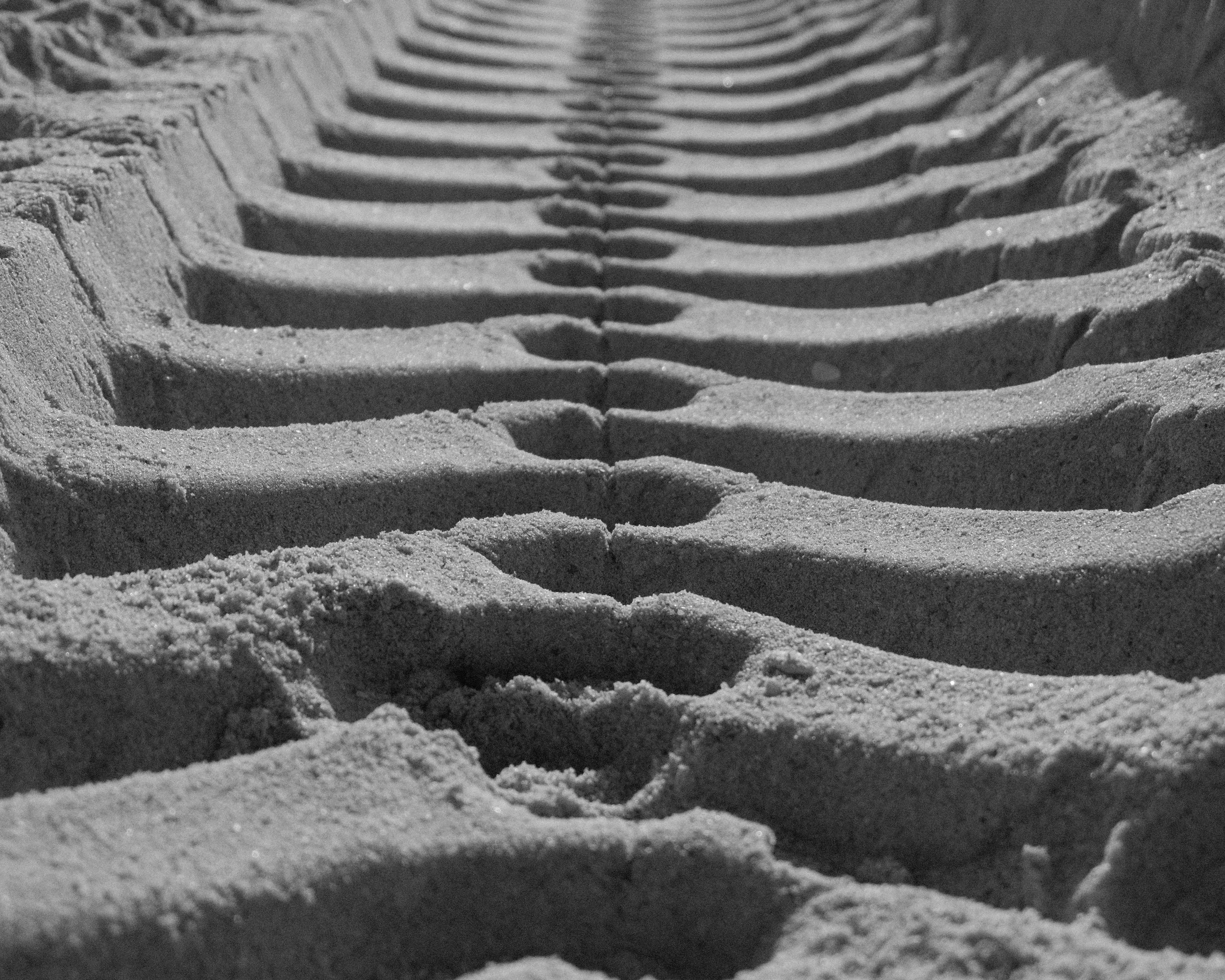 Close-up of tire tracks in sand on a beach, black and white photograph.
