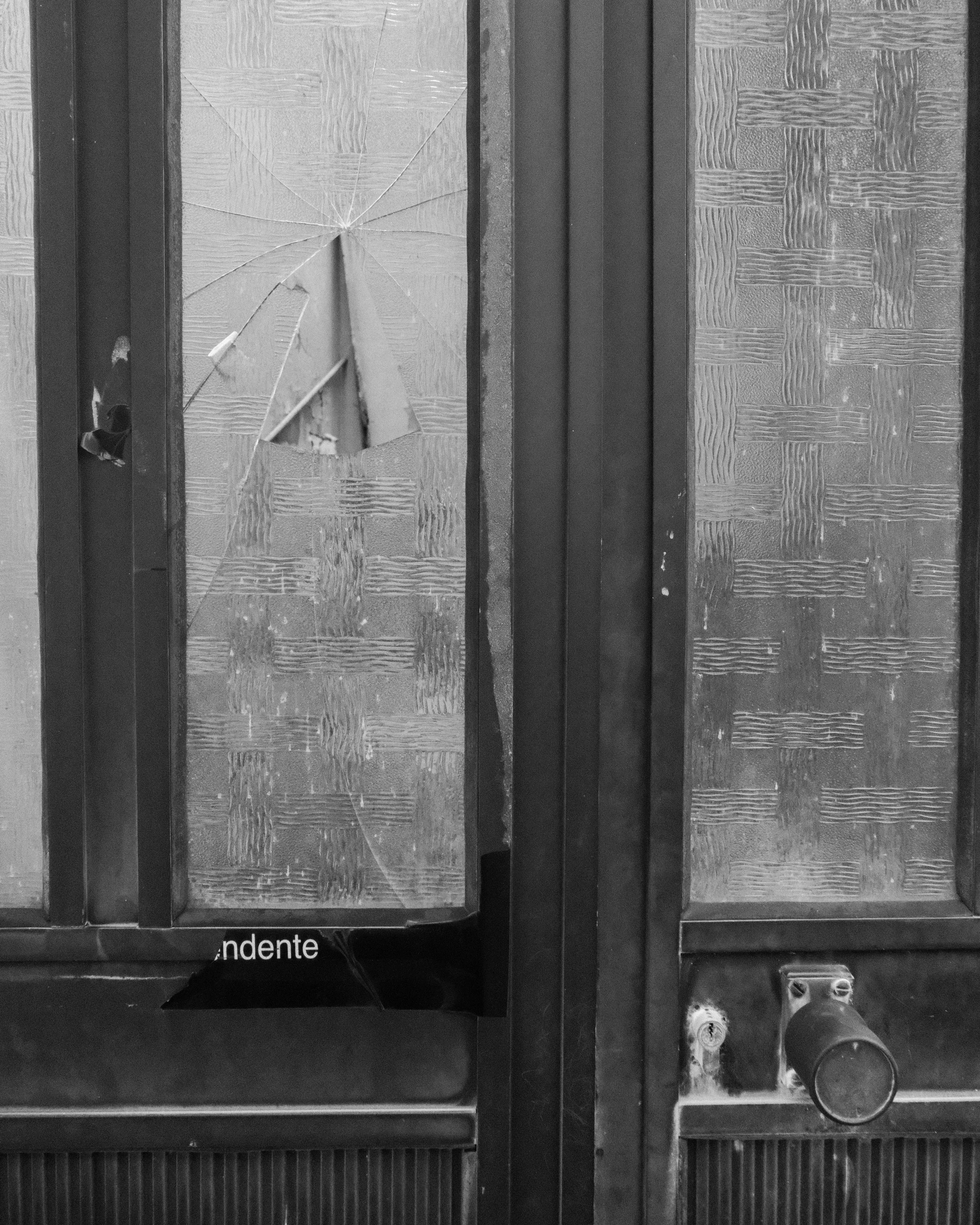A broken glass door with a large hole and cracks, part of a wooden cabinet or fridge with textured glass panels.