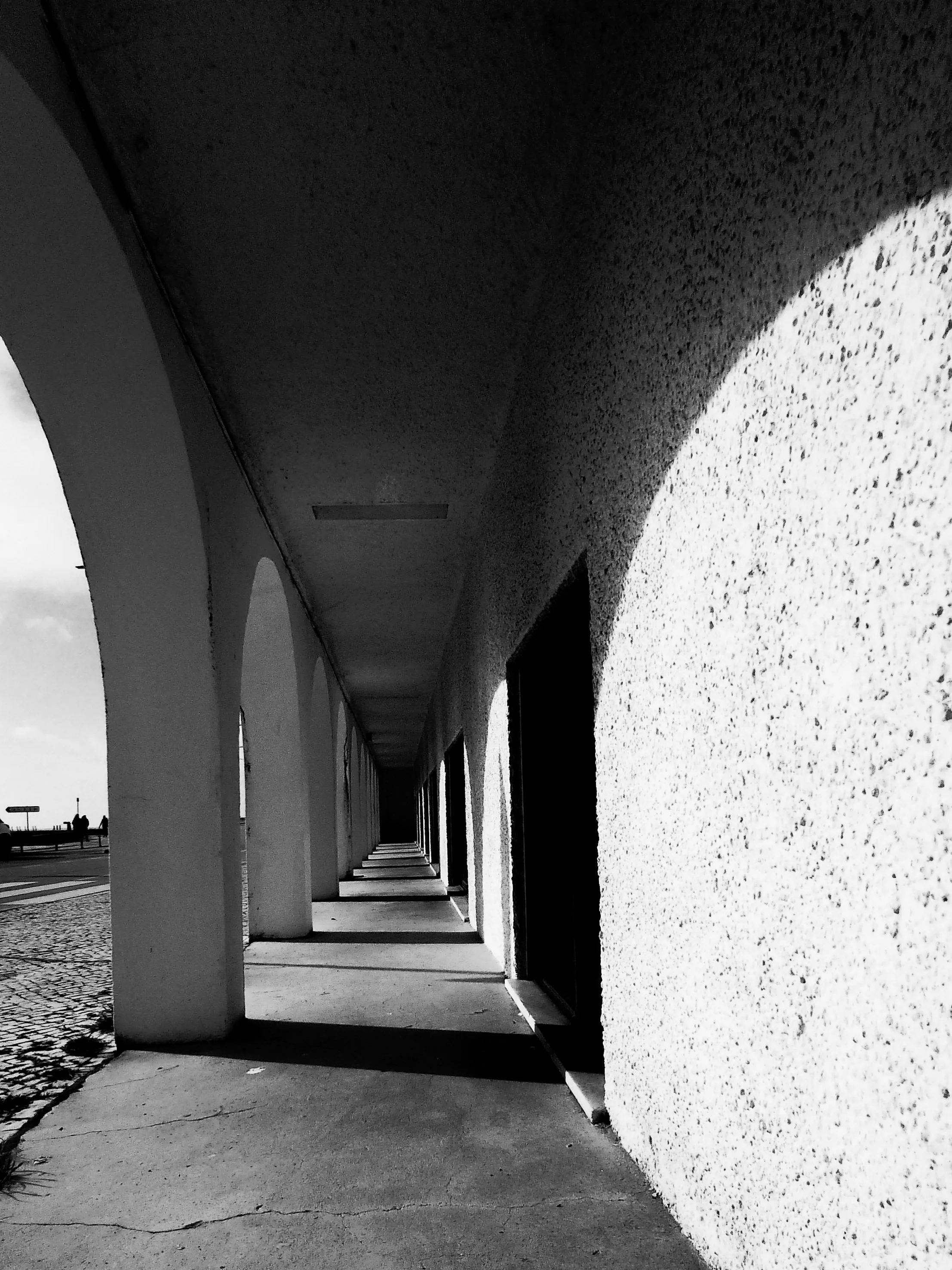 A black and white photo of a long outside arcade with arches along one side, casting shadows on the sidewalk.