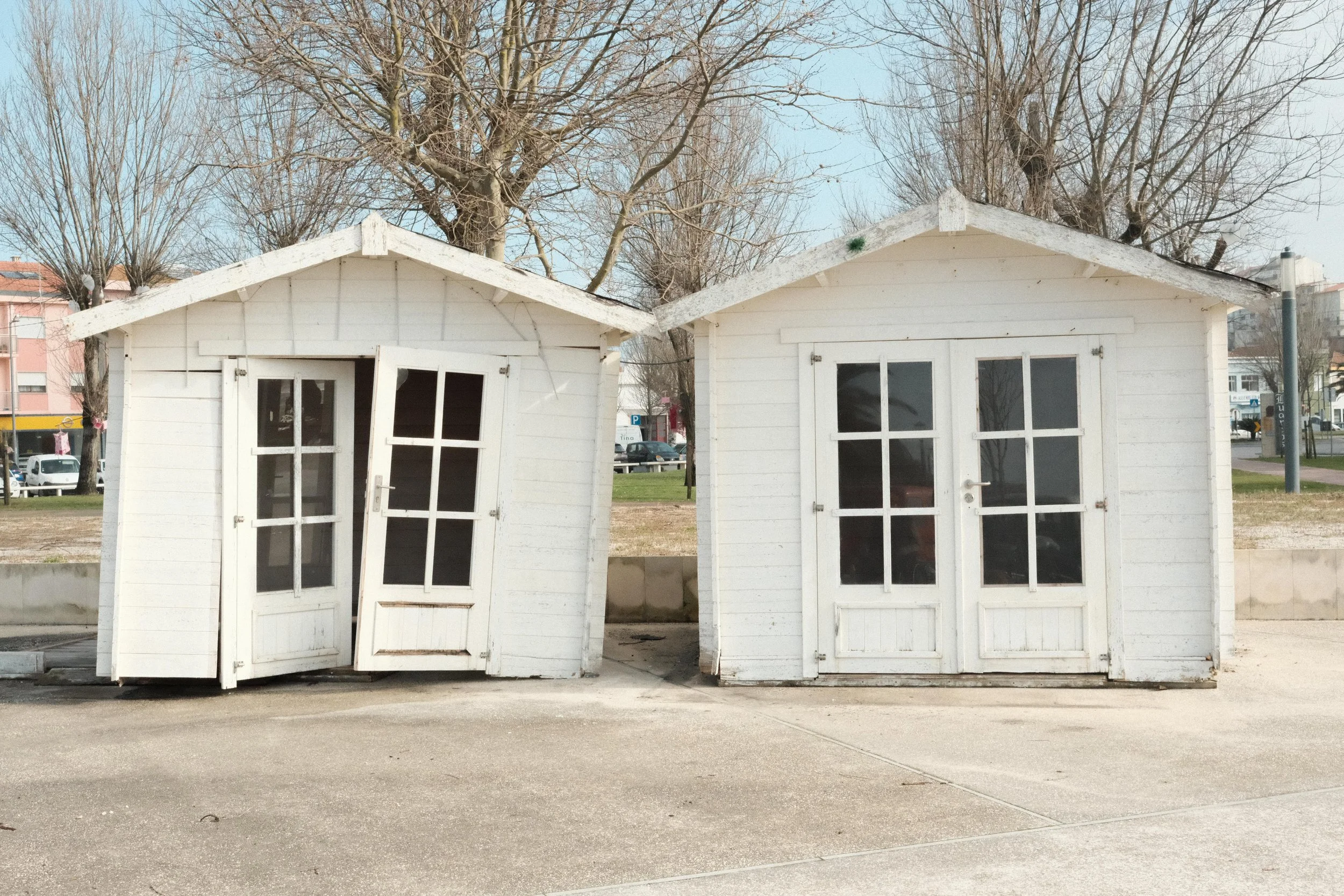 Two small white wooden houses with glass-paneled doors, one leaning slightly, are placed side by side on a paved surface outdoors. Behind them are leafless trees and a park area.