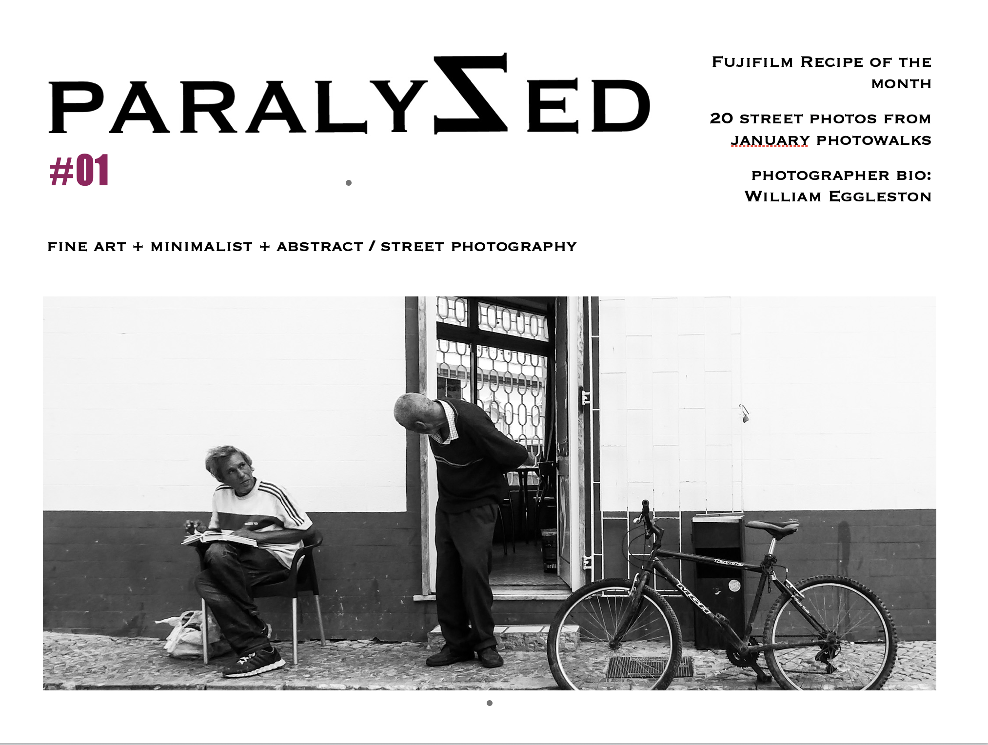 Black and white street scene with two men, one sitting on a chair reading a book and the other standing, talking to him, near a bicycle and a door.