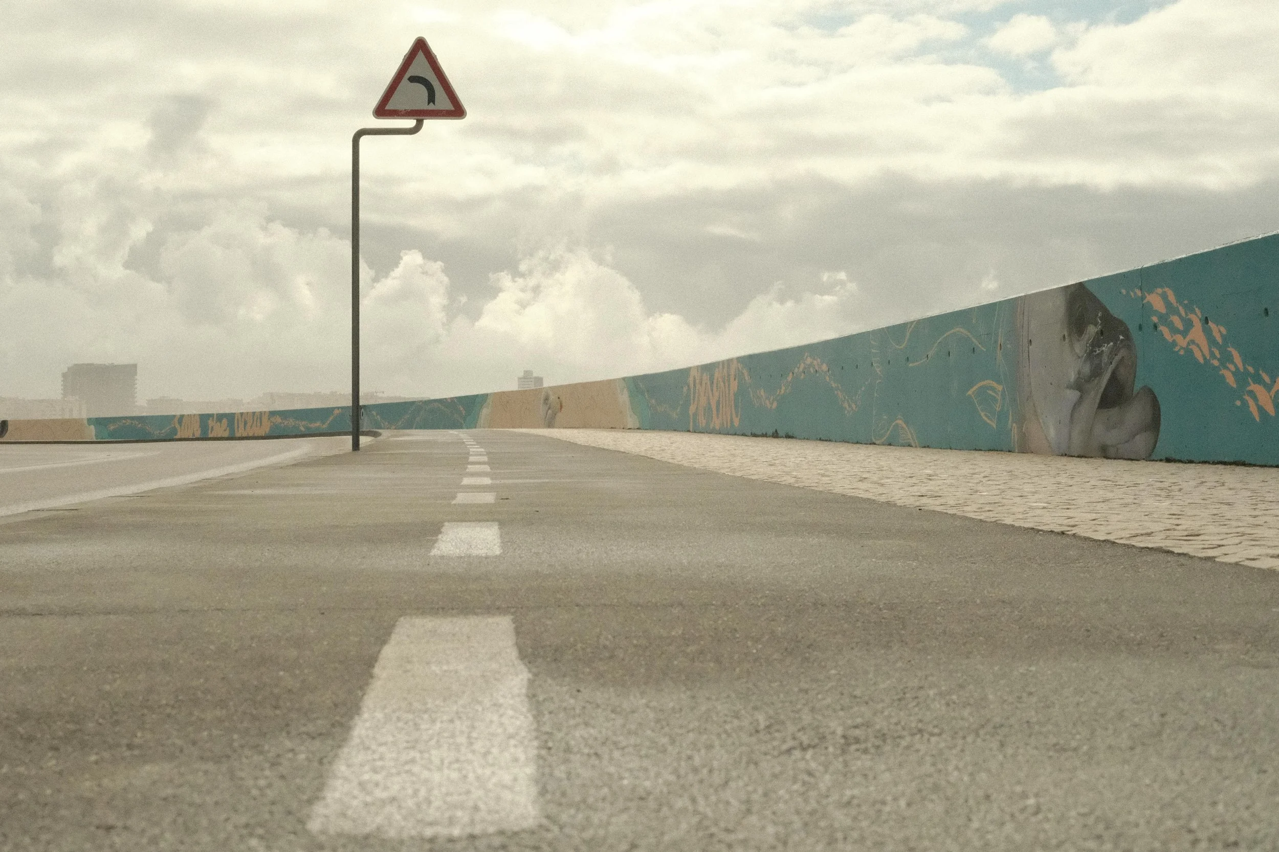 Empty sidewalk and road with a traffic sign indicating a left turn, along a colorful mural wall under cloudy sky.