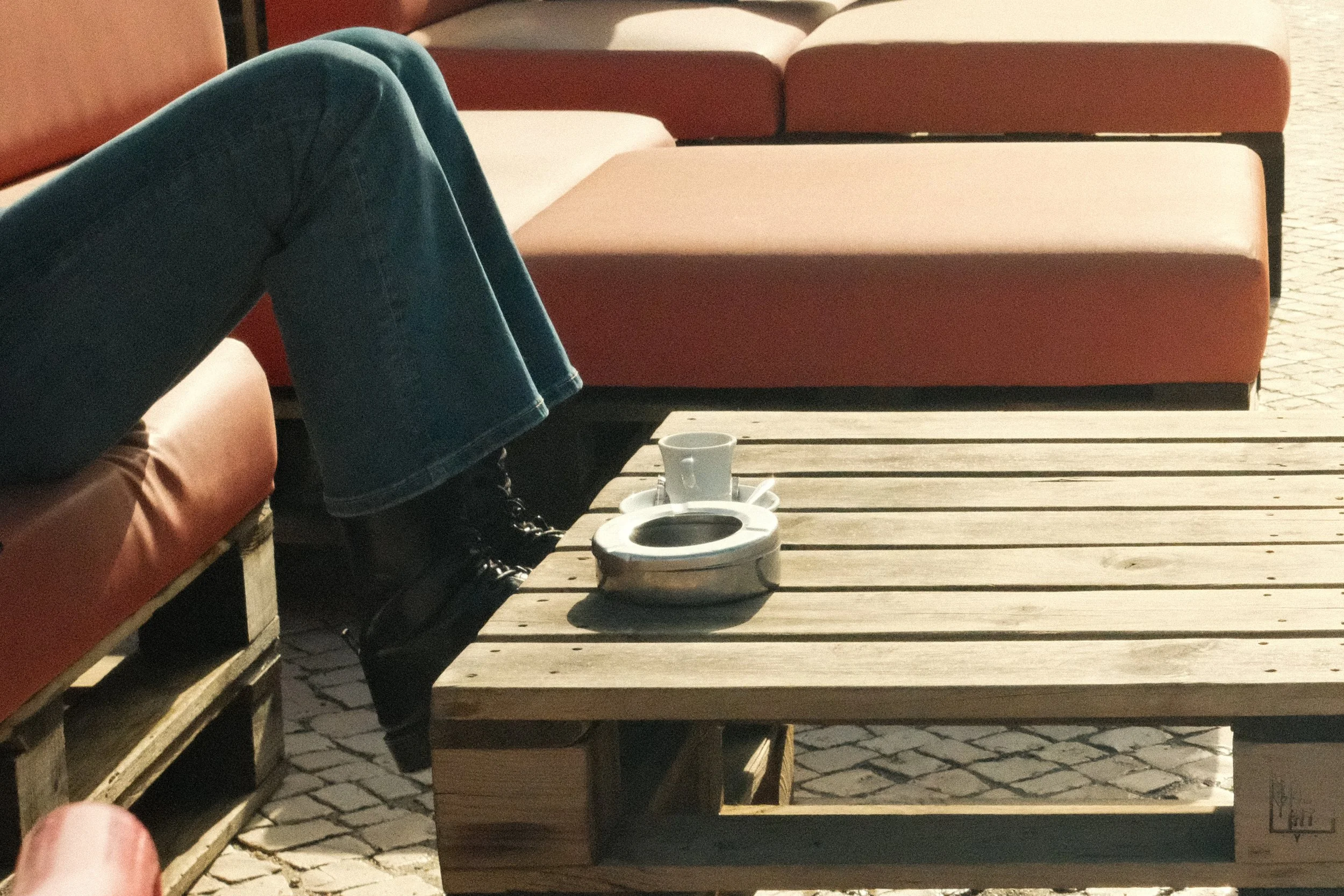 Person sitting on a sofa with legs crossed, coffee mug, and ashtray on wooden pallet table outside on a sunny day.
