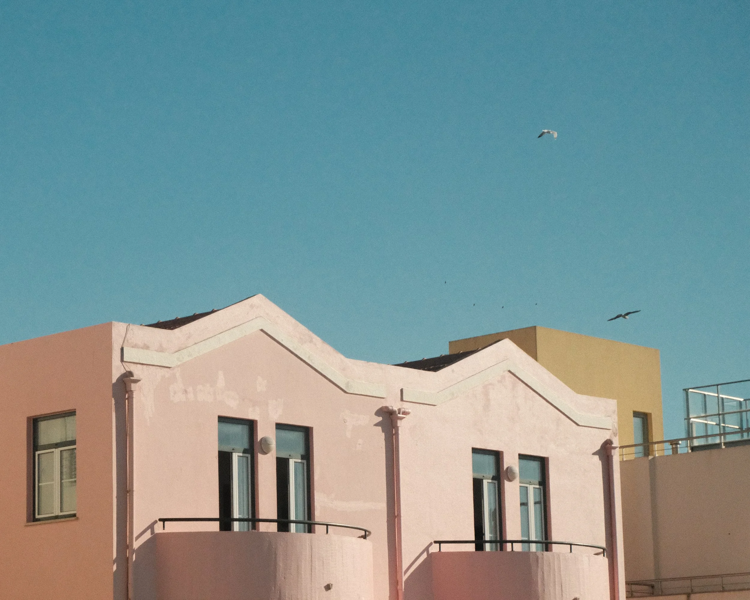 A pastel pink building with two balconies and large windows, against a clear blue sky with two seagulls flying.