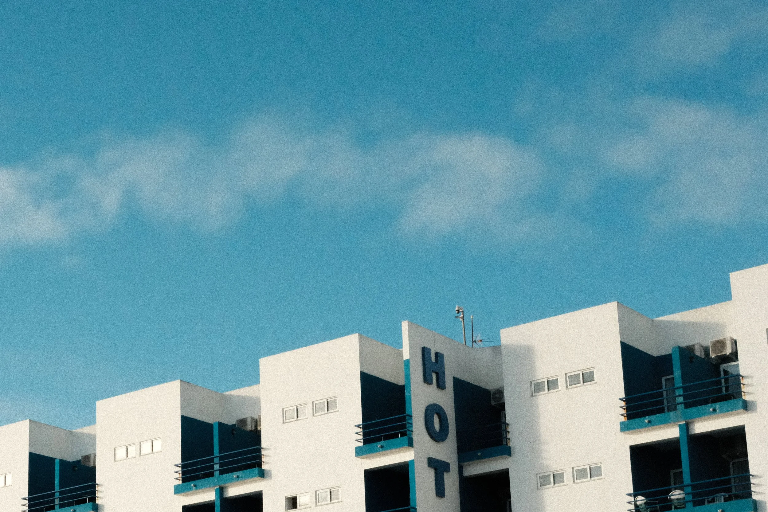 Part of a modern white hotel building with multiple balconies and a large vertical sign that reads 'HOT'. The building has blue accents and is set against a clear blue sky with a few clouds.