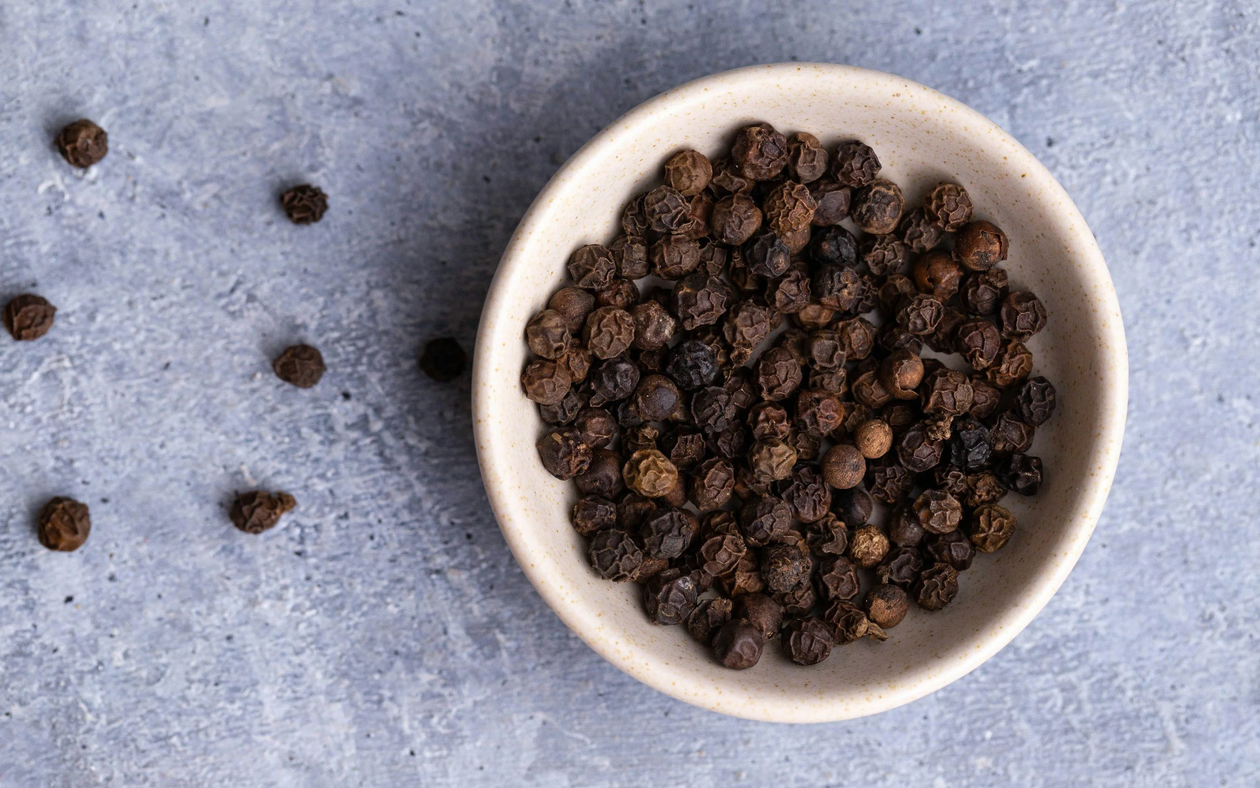 A white bowl filled with black peppercorns on a light gray textured background with some peppercorns scattered around.