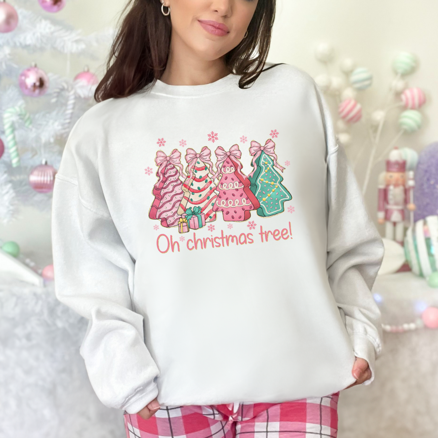 Woman wearing a white sweatshirt with Christmas tree designs and the words "Oh christmas tree!" in front of a decorated Christmas tree and holiday decorations.