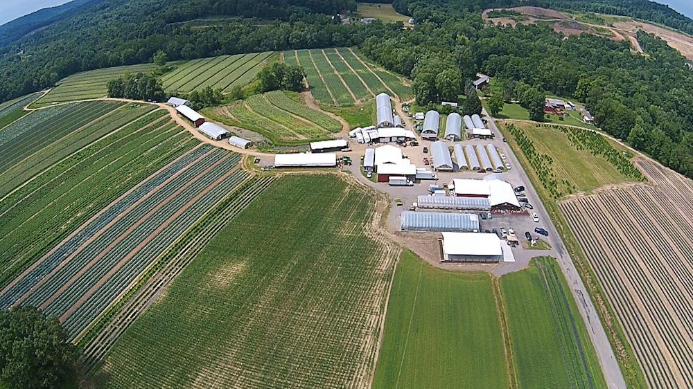 Aerial view of Yarnick's farm with fields, greenhouses, and farm buildings surrounded by hills and trees.
