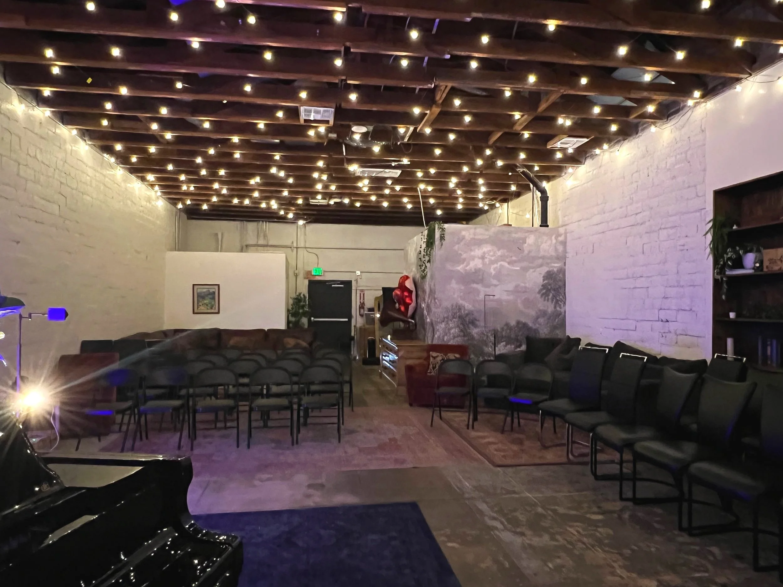 Interior of a room with string lights, white brick walls, a black piano in the foreground, and rows of empty chairs. The room has a cozy ambiance with some plush seating and a mural on one wall.