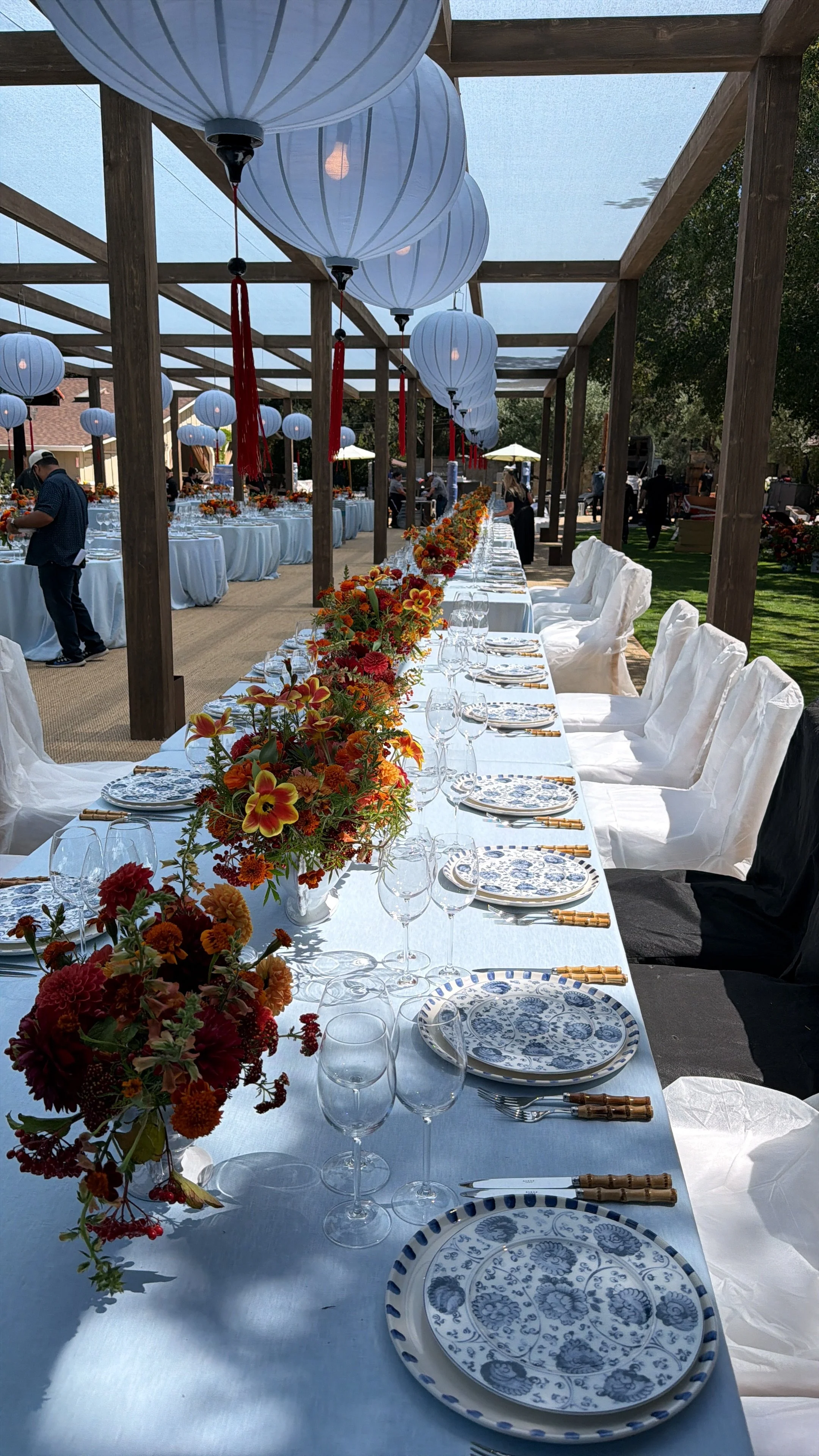 Outdoor banquet table with floral centerpieces, blue-and-white place settings and hanging lanterns.