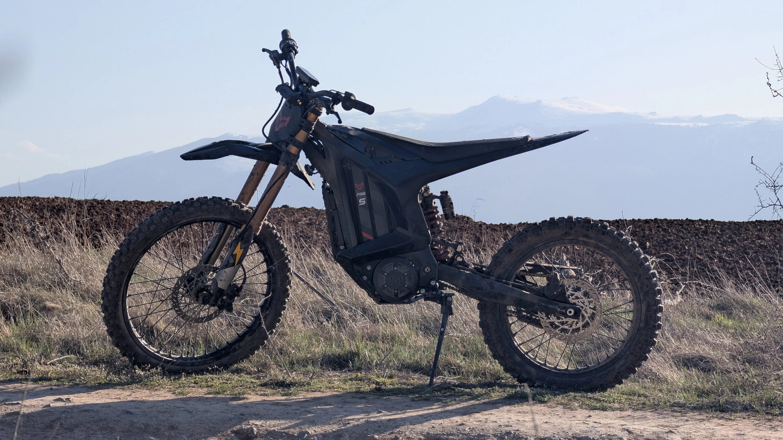 An electric dirt bike with a black body and off-road tires parked on a dirt path with mountains in the background.
