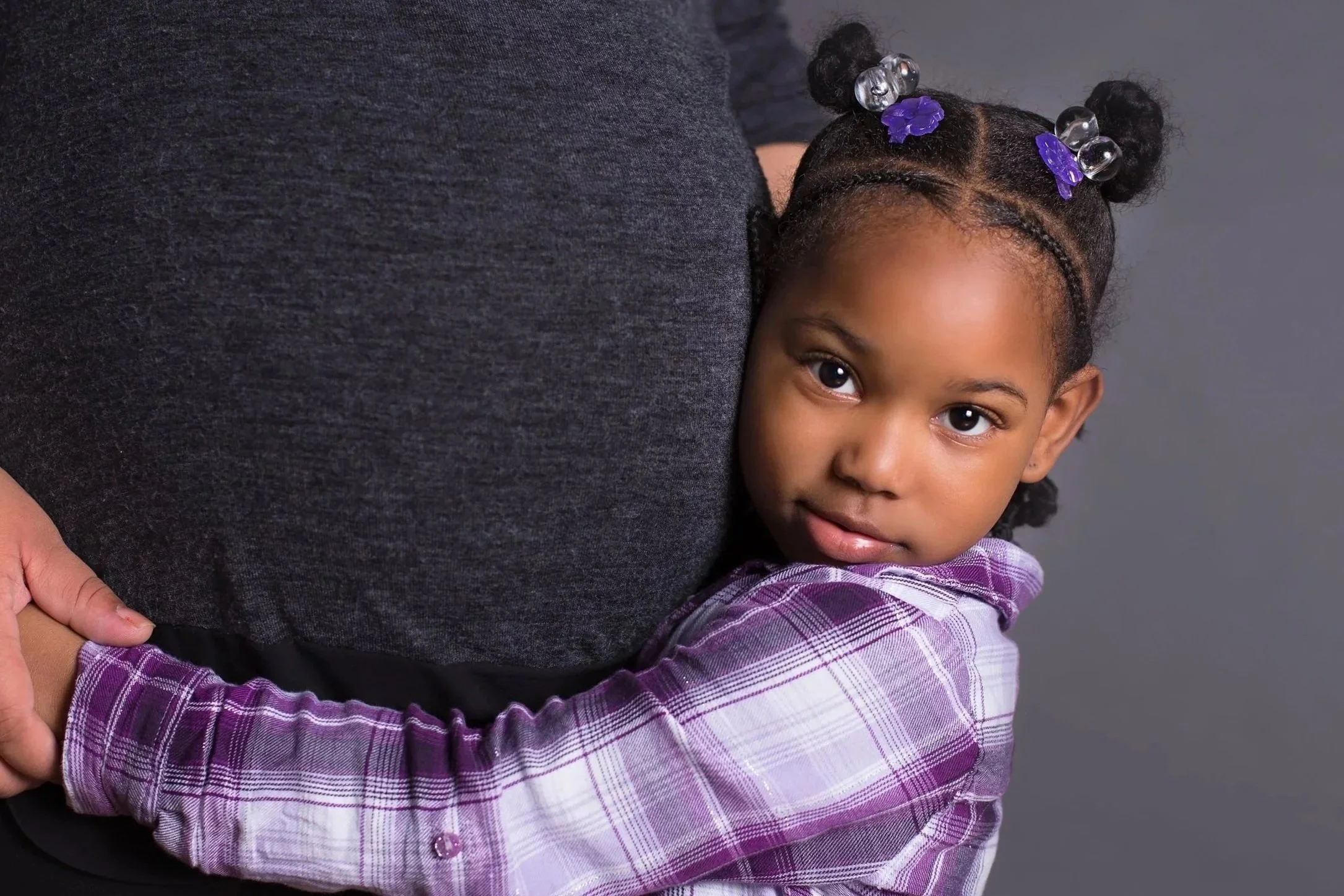 Young girl with braided hair, purple bows, and beads, hugging an adult wearing a dark gray shirt against a gray background.