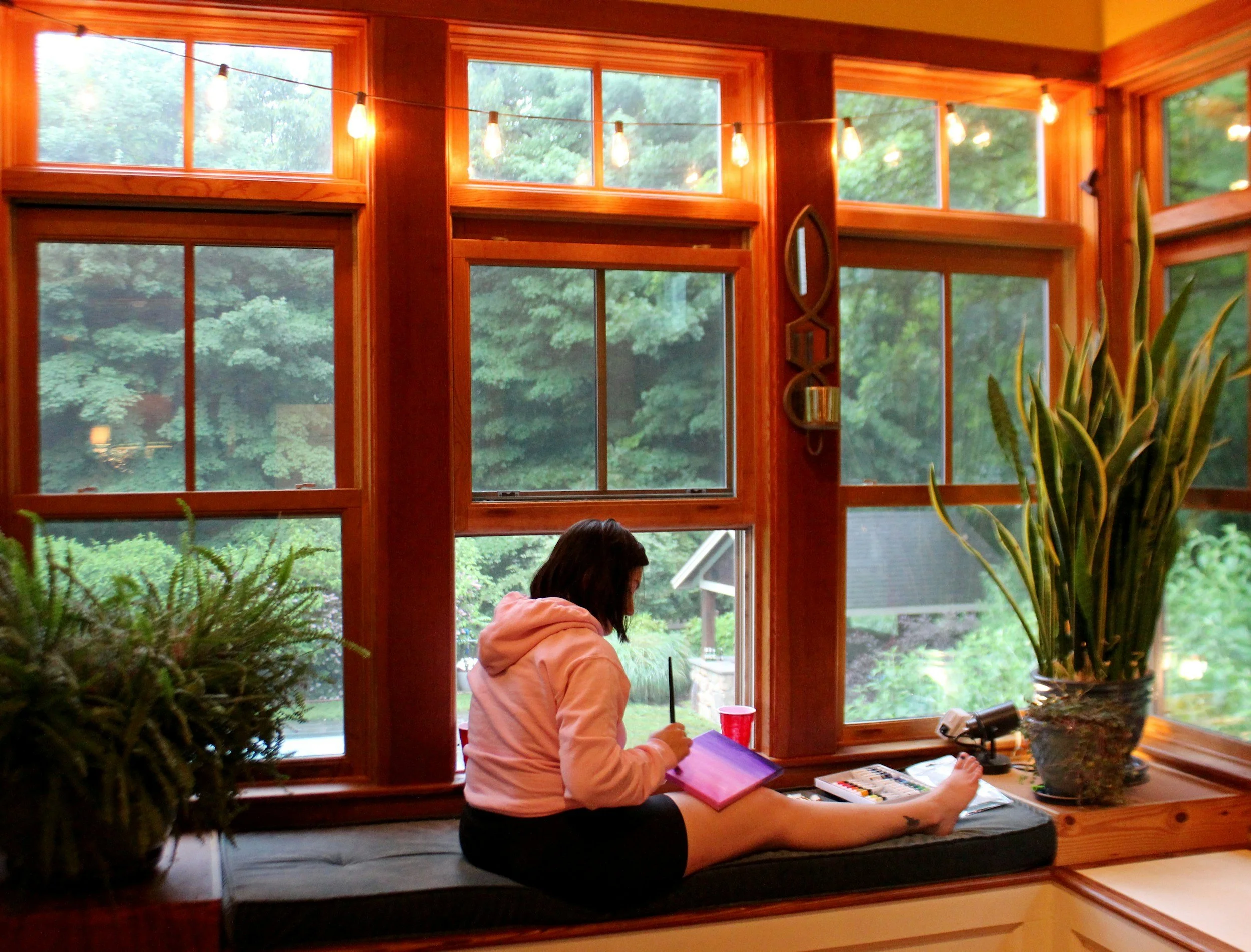 a person sitting on a black cushioned  window seat within a wooden alcove painting in between plants.