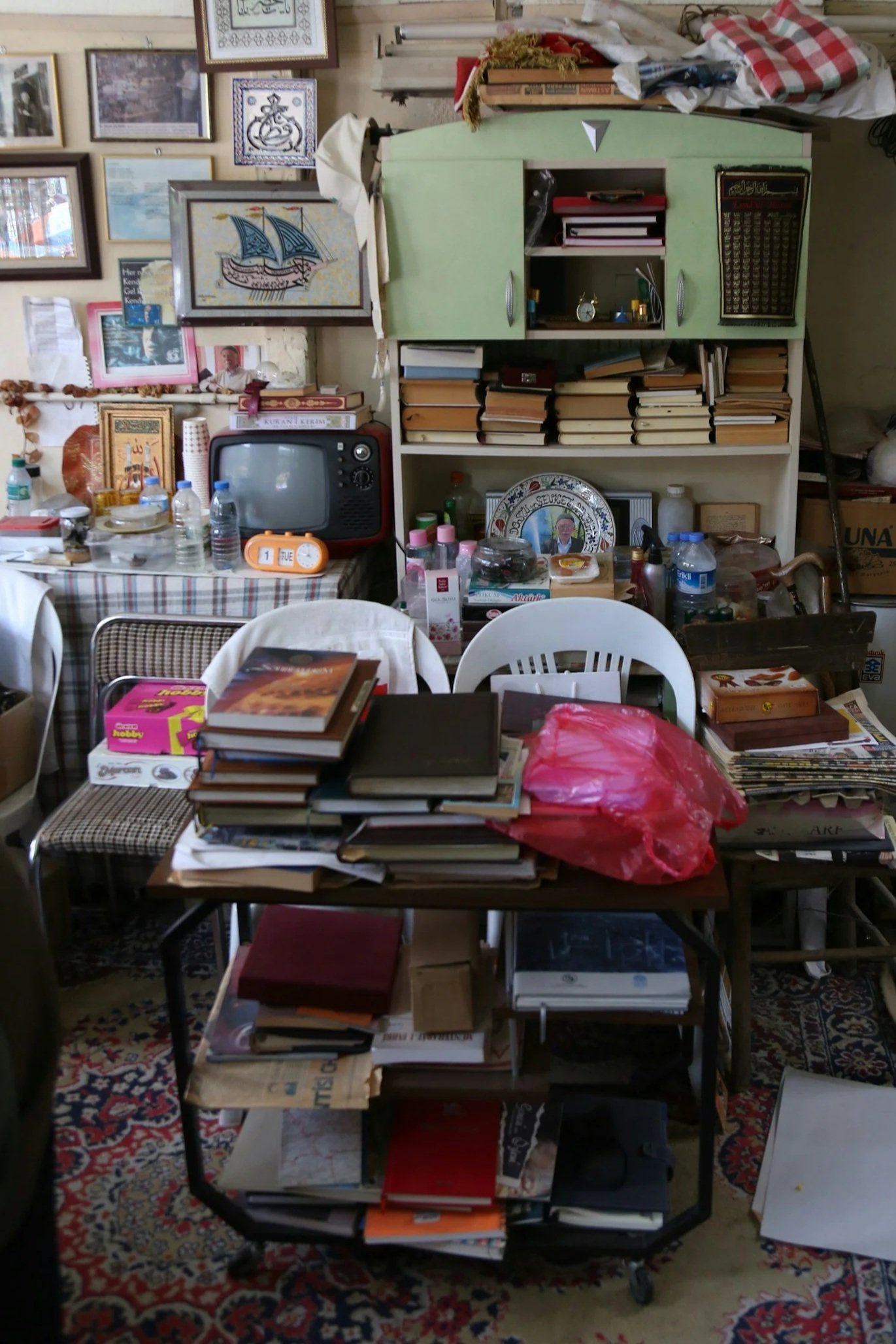 A room filled with books everywhere, on a drinks trolley, in open shelves, piled on chairs. Samson Projects can help with organising this space. The walls are full with all kinds of pictures and frames. The floor has a large ornate rug.