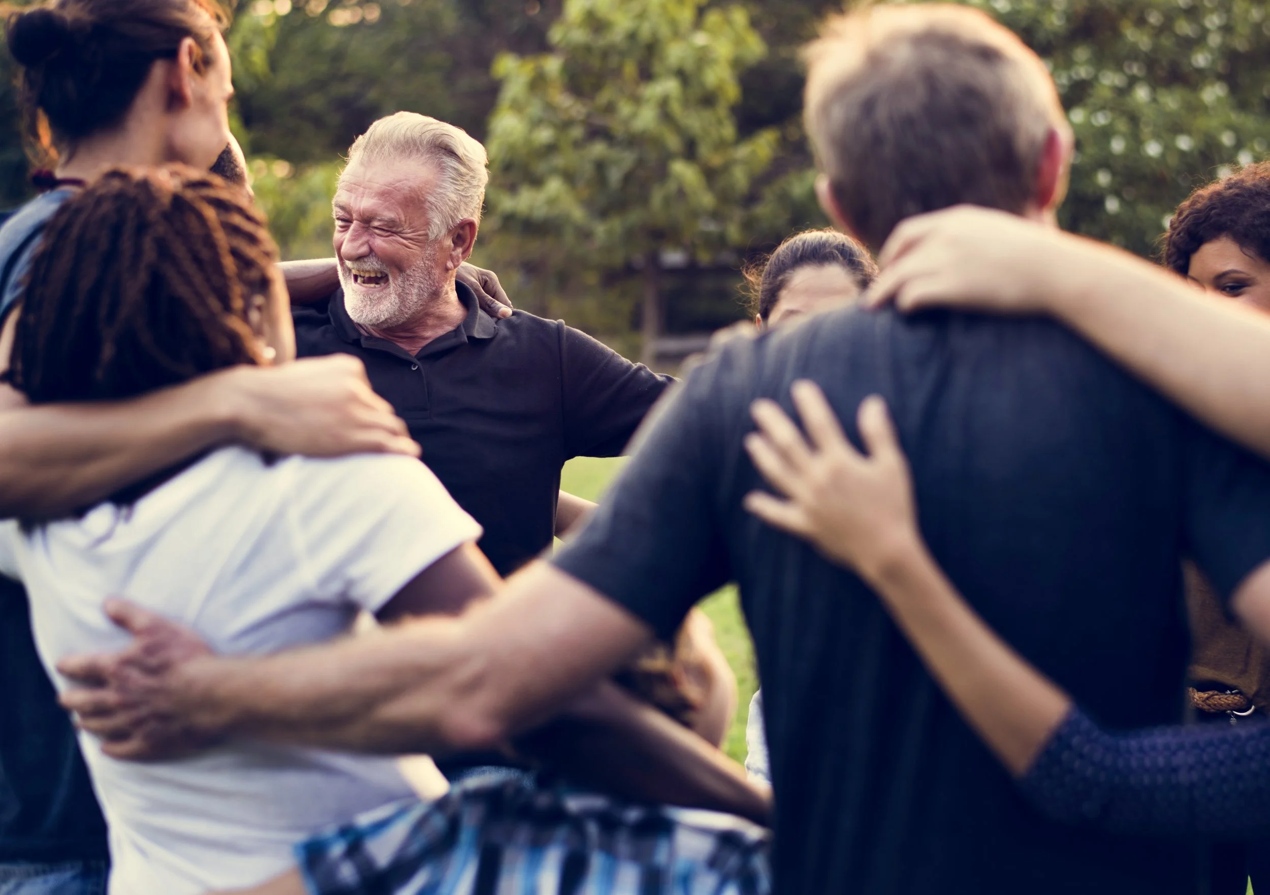 A diverse group of people embraces and smiles in a circle outdoors, enjoying a moment of connection and happiness.