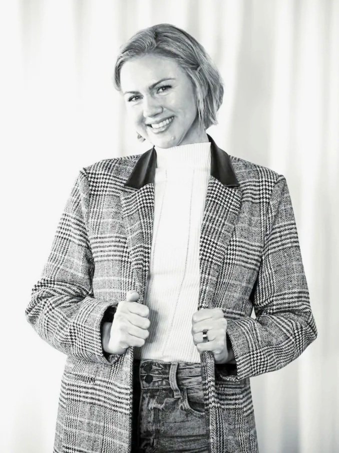 A woman with short hair smiling, wearing a plaid blazer with leather collar, white turtleneck, and jeans, standing in front of light-colored curtains.