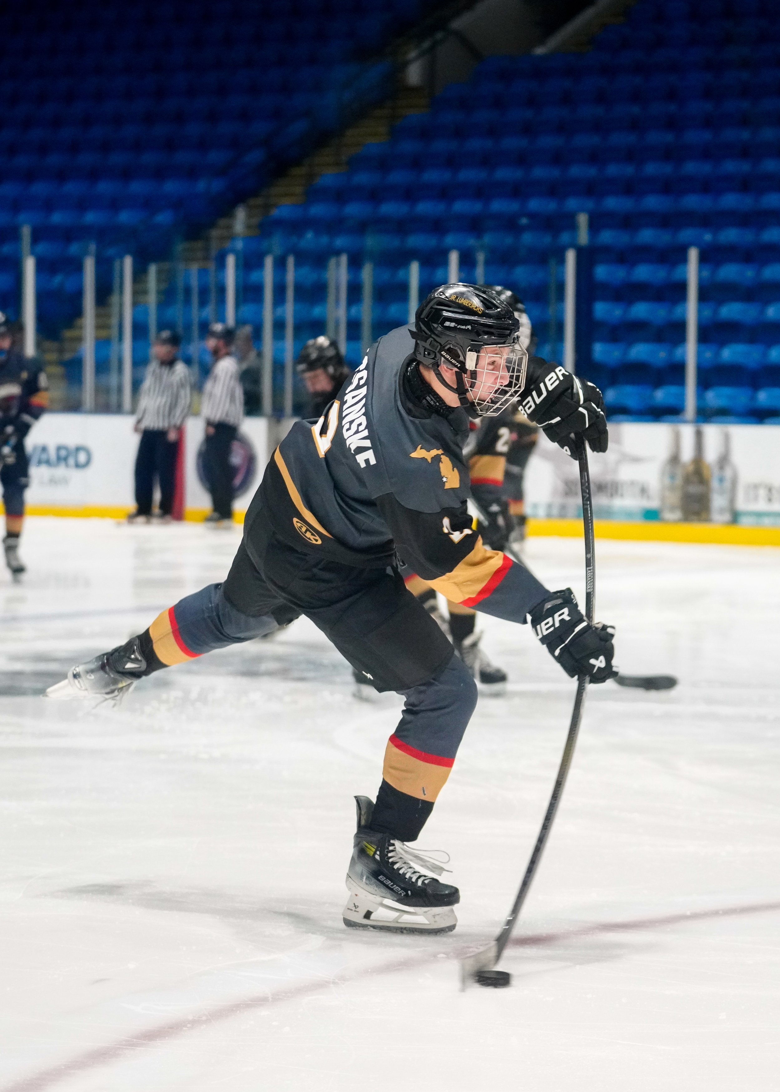 Hockey player in black and gold uniform on ice, taking a shot with a hockey stick on an indoor rink, with referees and spectators in the background.