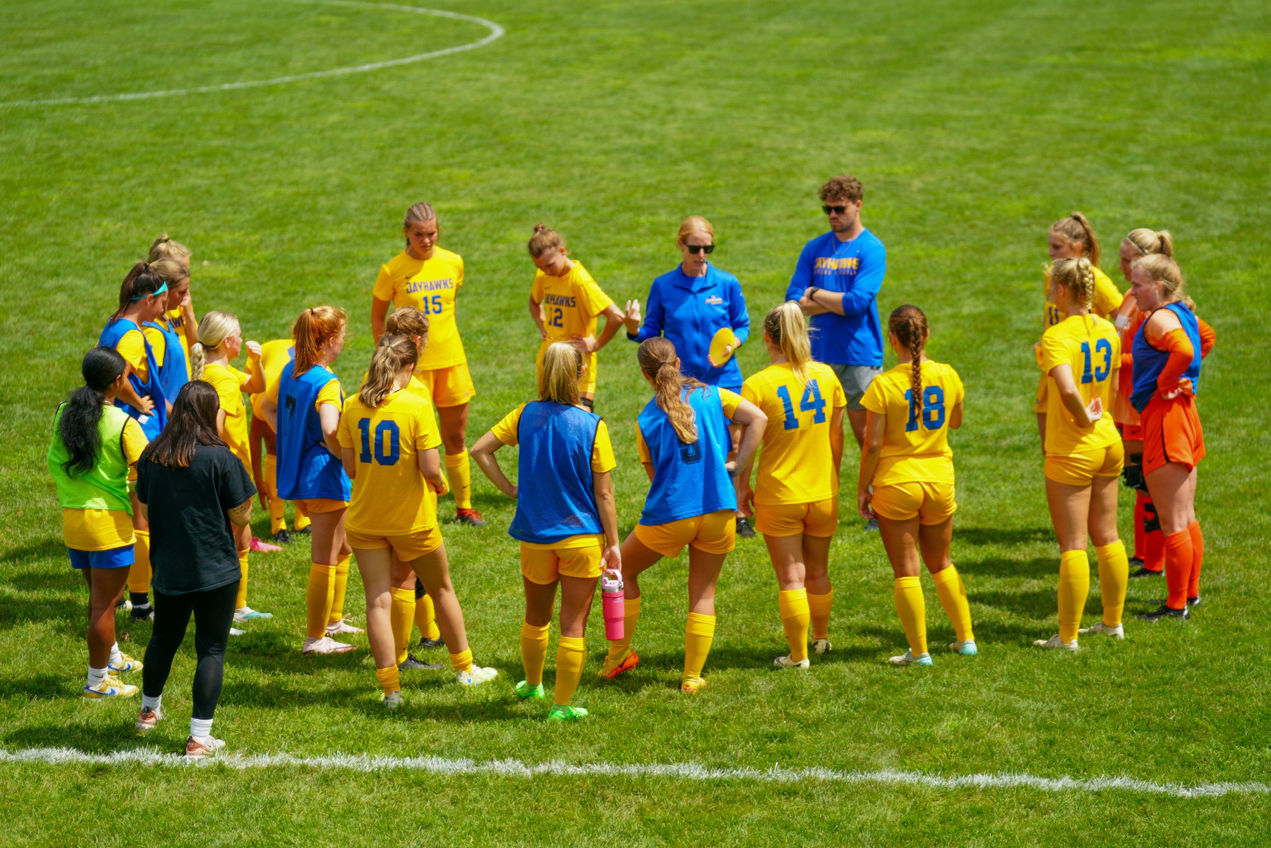 Women's soccer team huddling on a grass field wearing yellow jerseys and blue pinnies, with coaches giving instructions.