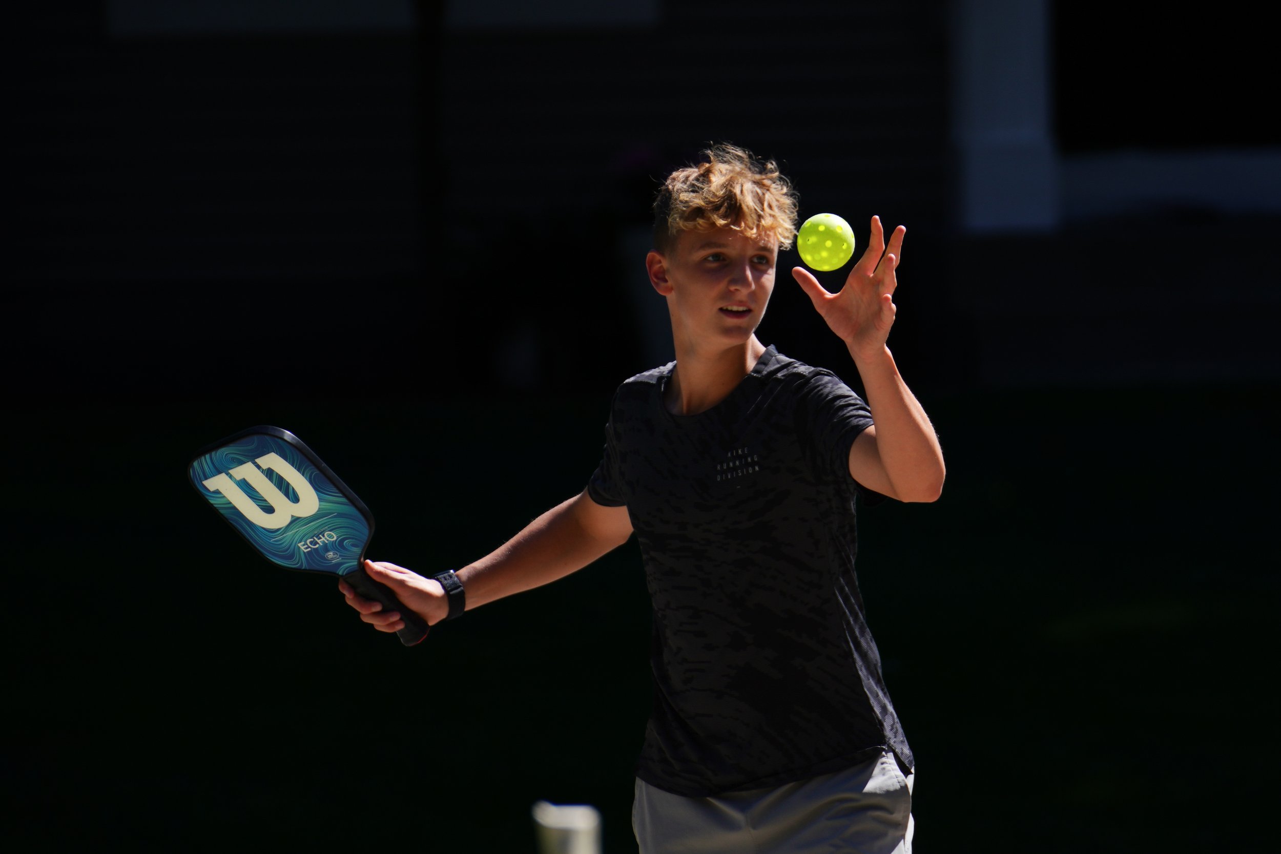 Person playing pickleball, holding a paddle and a green perforated ball, outdoors.