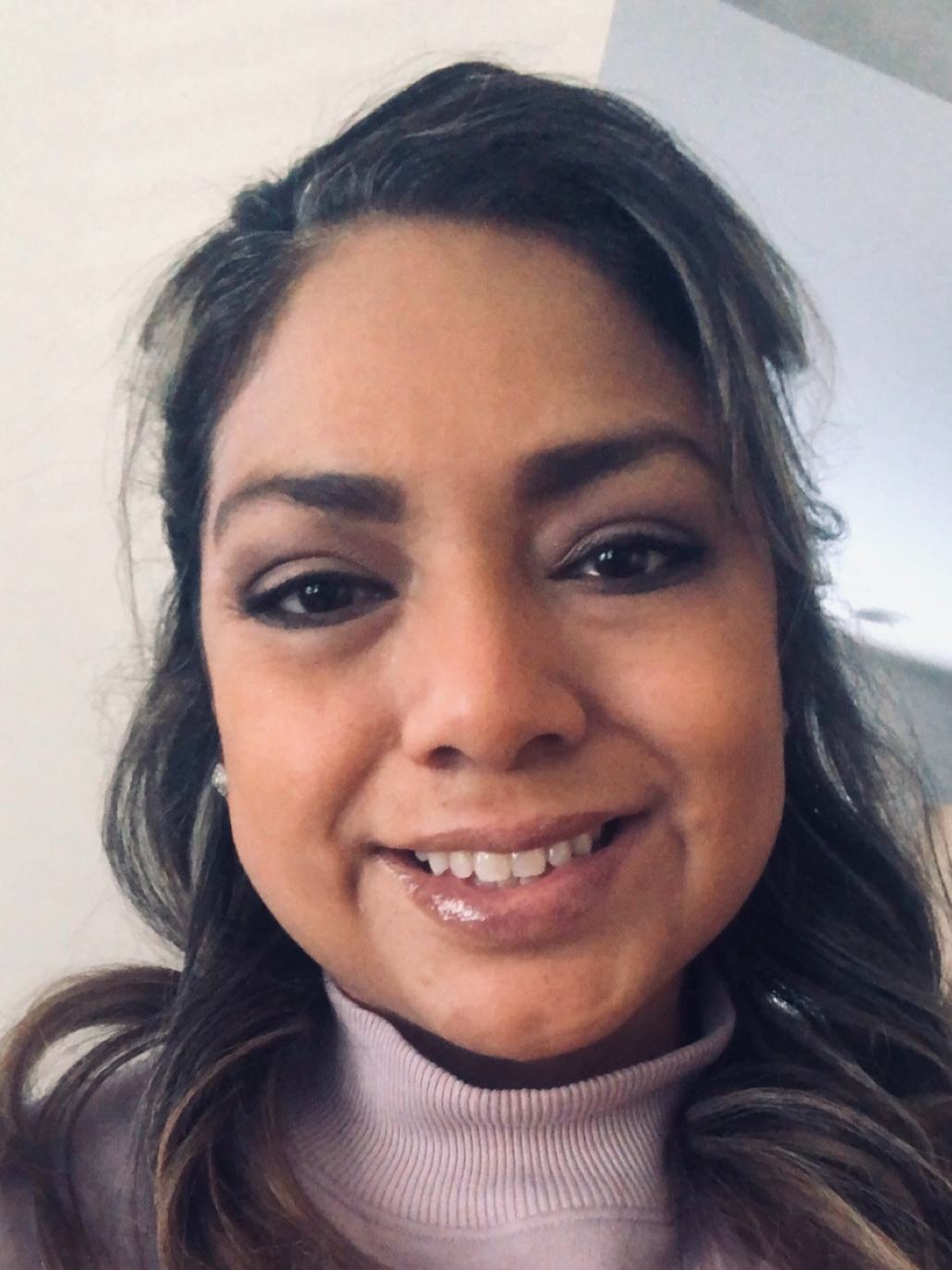 Close-up of a smiling woman with long, wavy dark hair, wearing a light-colored turtleneck sweater, indoors with plain walls in the background.