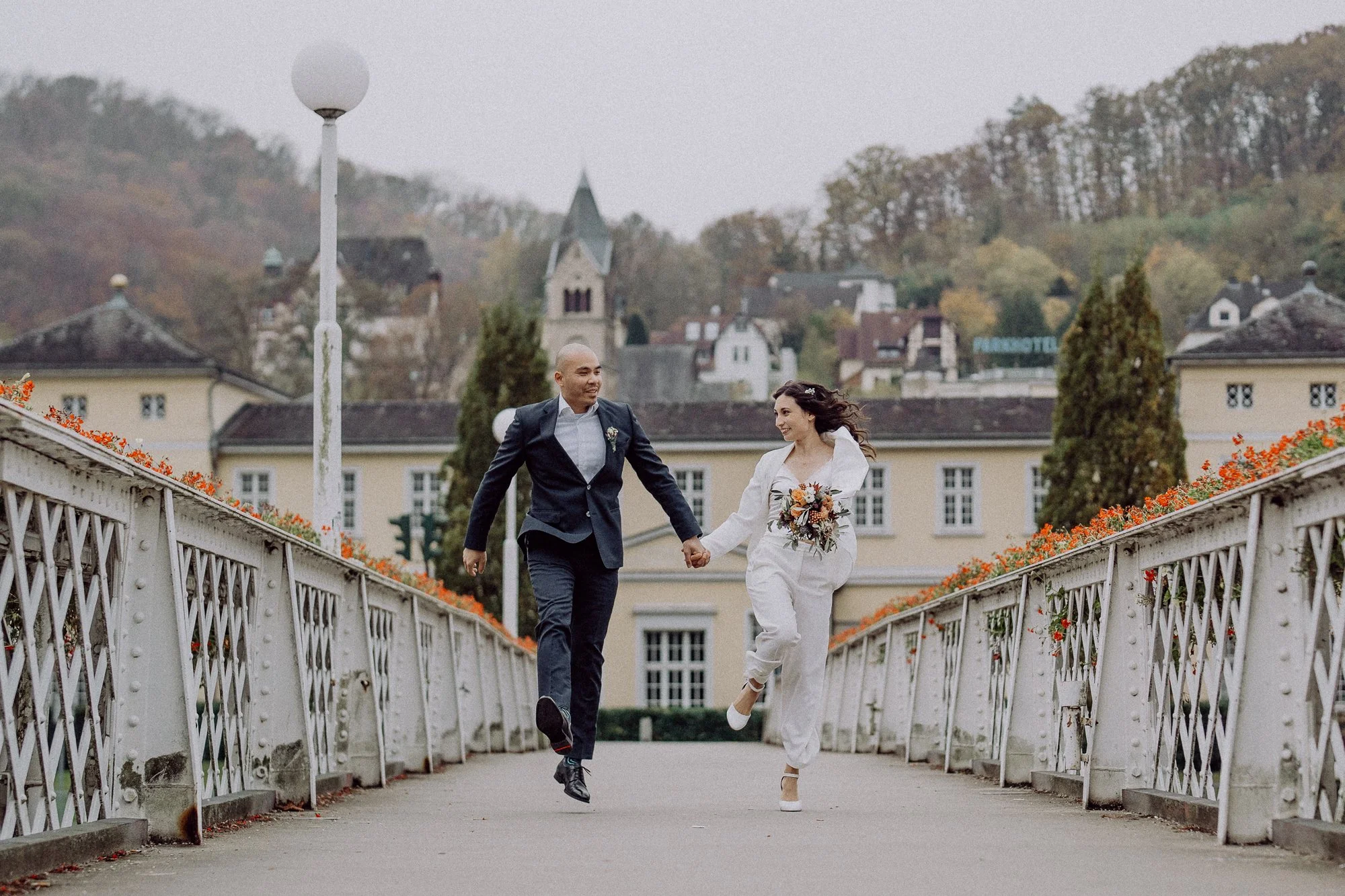 Ein Paar läuft über eine Brücke Hand in Hand. Der Mann trägt einen Anzug, die Frau hält einen Blumenstrauß. Im Hintergrund sind Gebäude und Herbstbäume zu sehen.