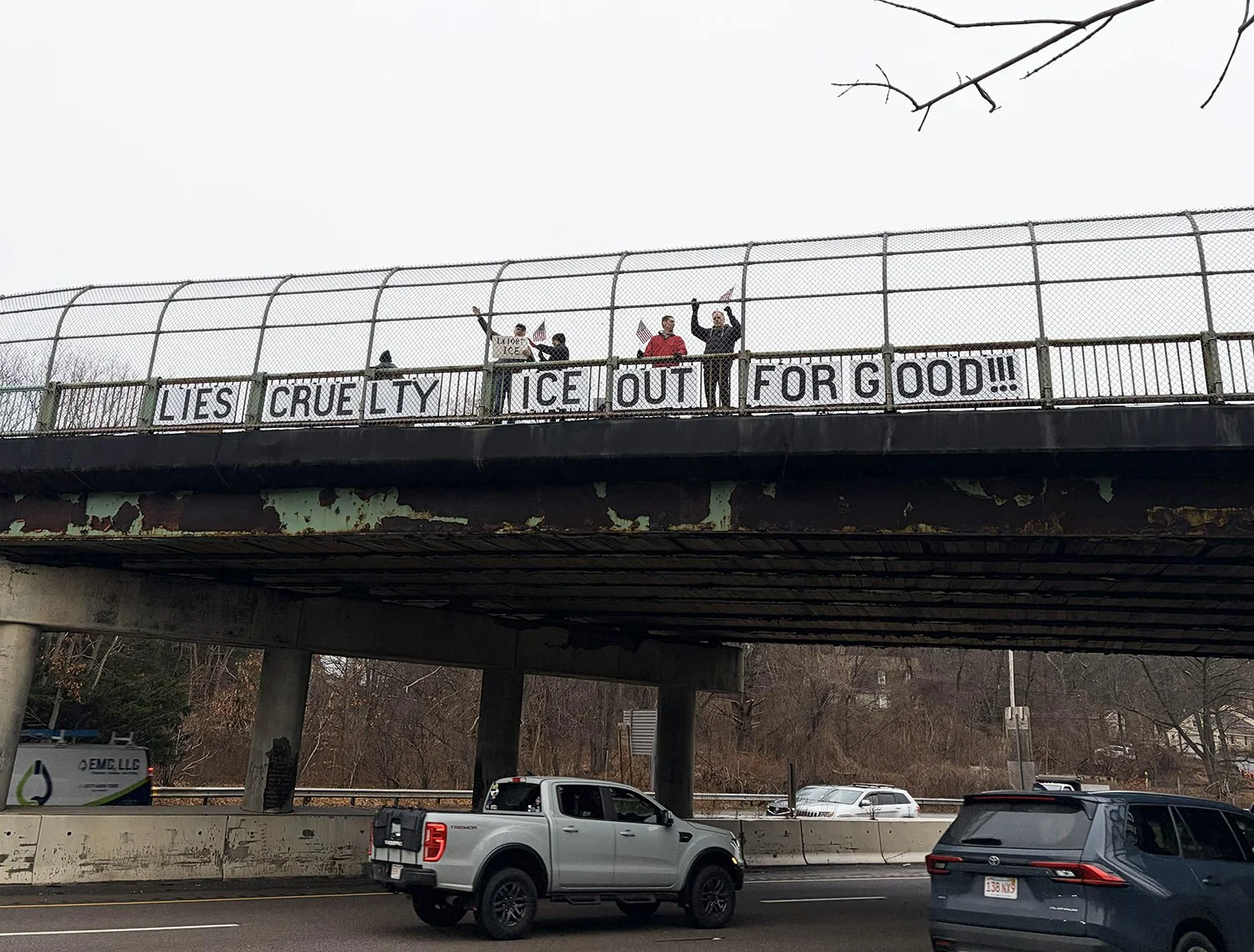 highway overpass in Waltham with signs reading "Lies, Cruelty" and "ICE Out For Good"
