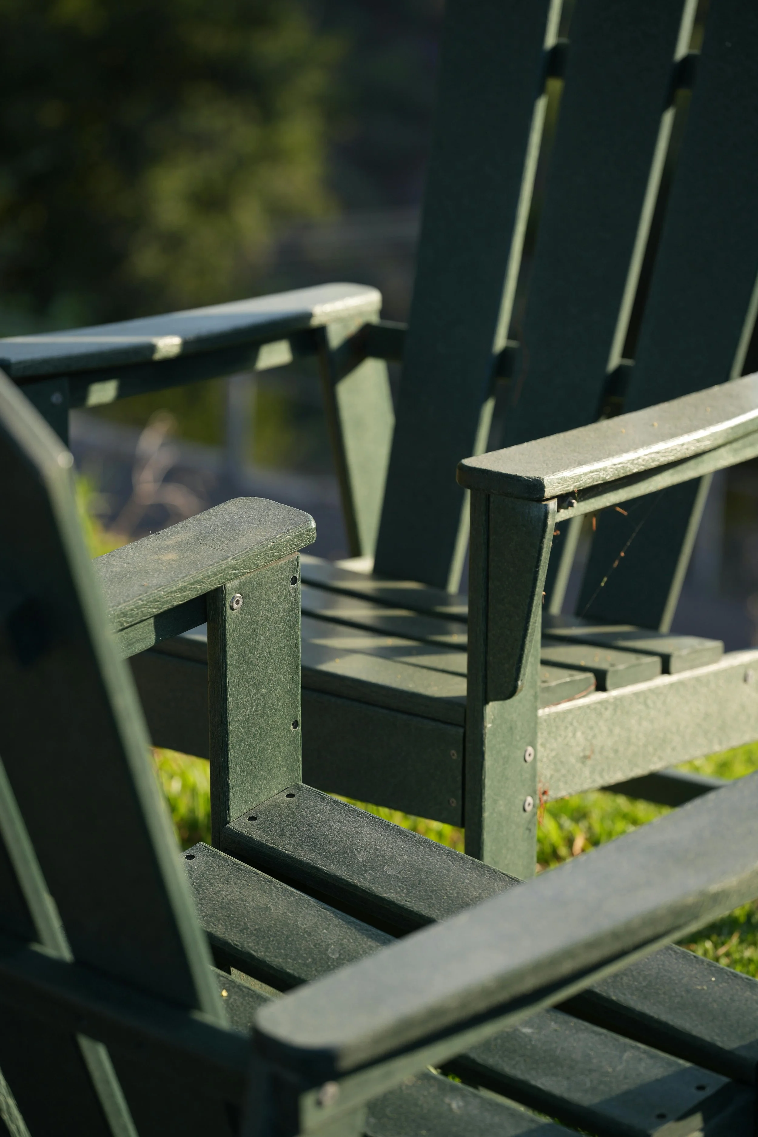 Close-up of green wooden outdoor Adirondack chairs on grass with sunlight.