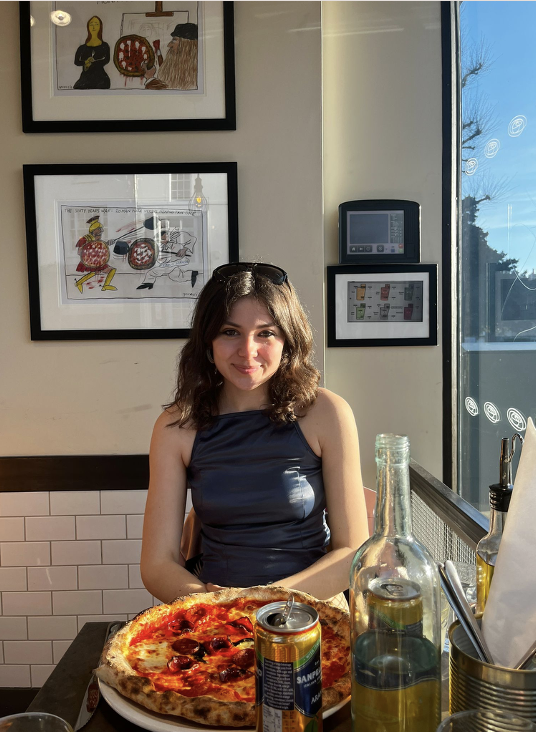 Young woman sitting at a table in a restaurant with a pizza, two bottles, a can of soda, and utensils, with framed artwork on the wall and a window showing bright daylight outside.