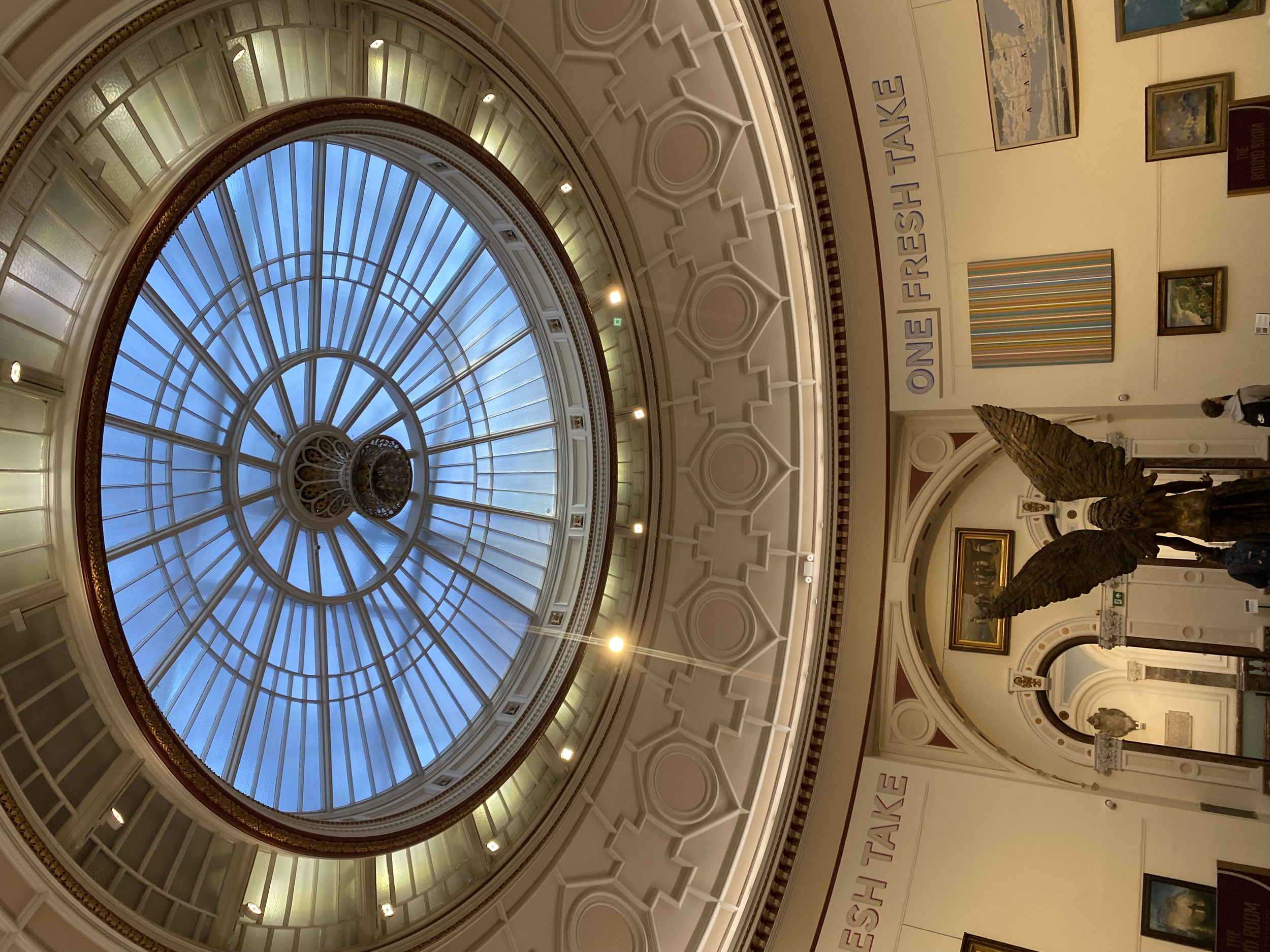 Interior of a grand building with a circular glass dome ceiling, art displays, and a large eagle sculpture.
