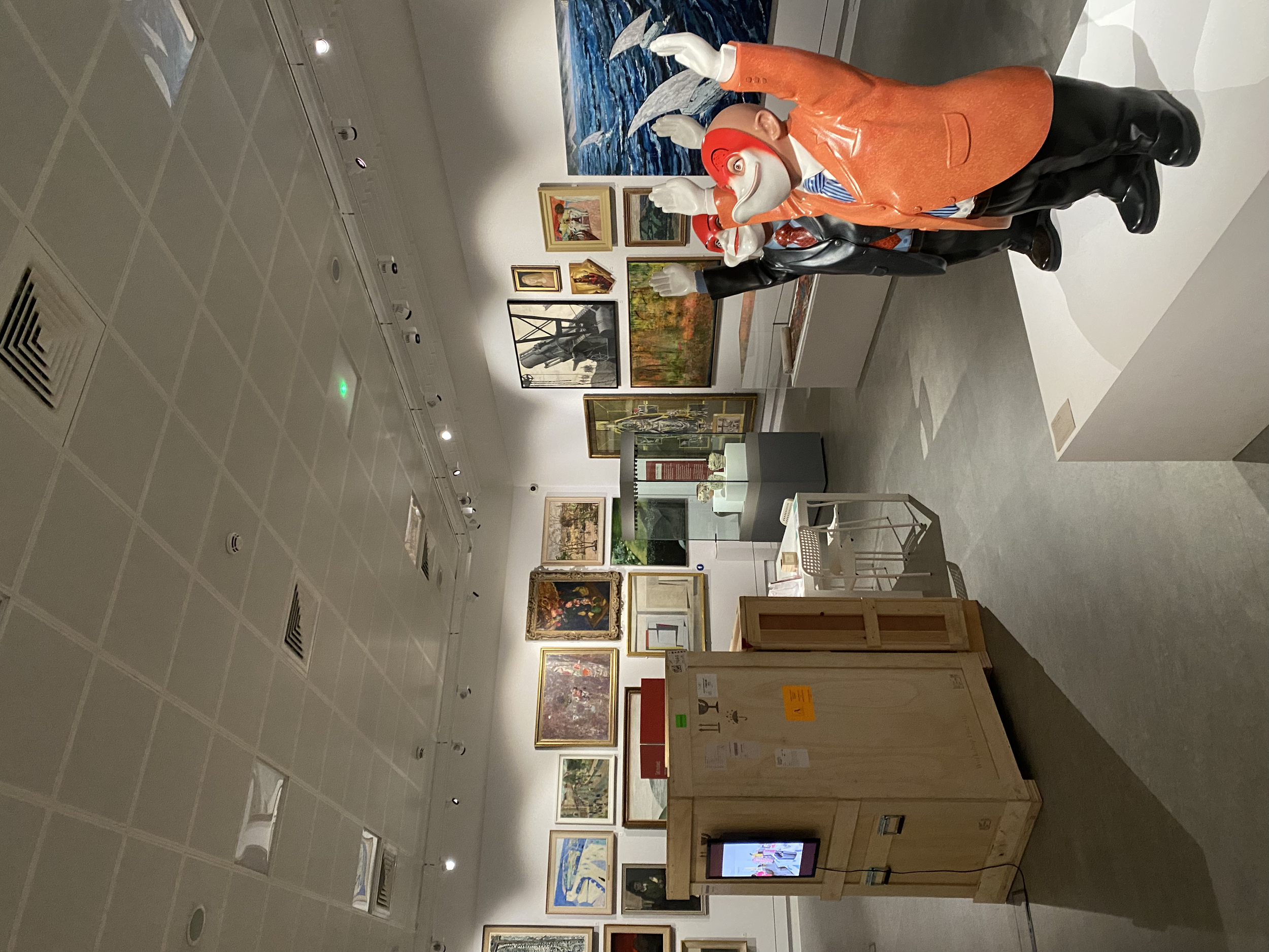 Gallery room with various framed artworks on the walls and a large, colorful sculpture of a person wearing an orange jacket and hat in the foreground. There is also a crate with a monitor showing a video.