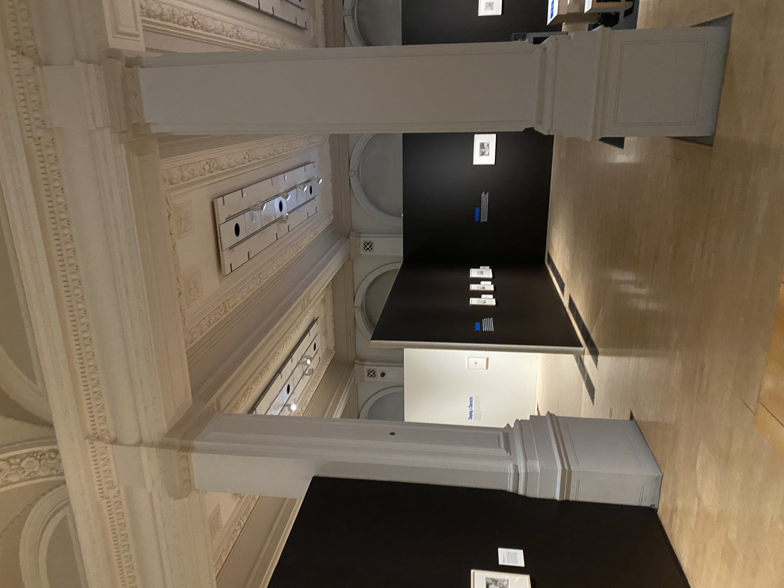 Interior of an art gallery with ornate columns, wooden floors, and black display walls featuring framed artwork.