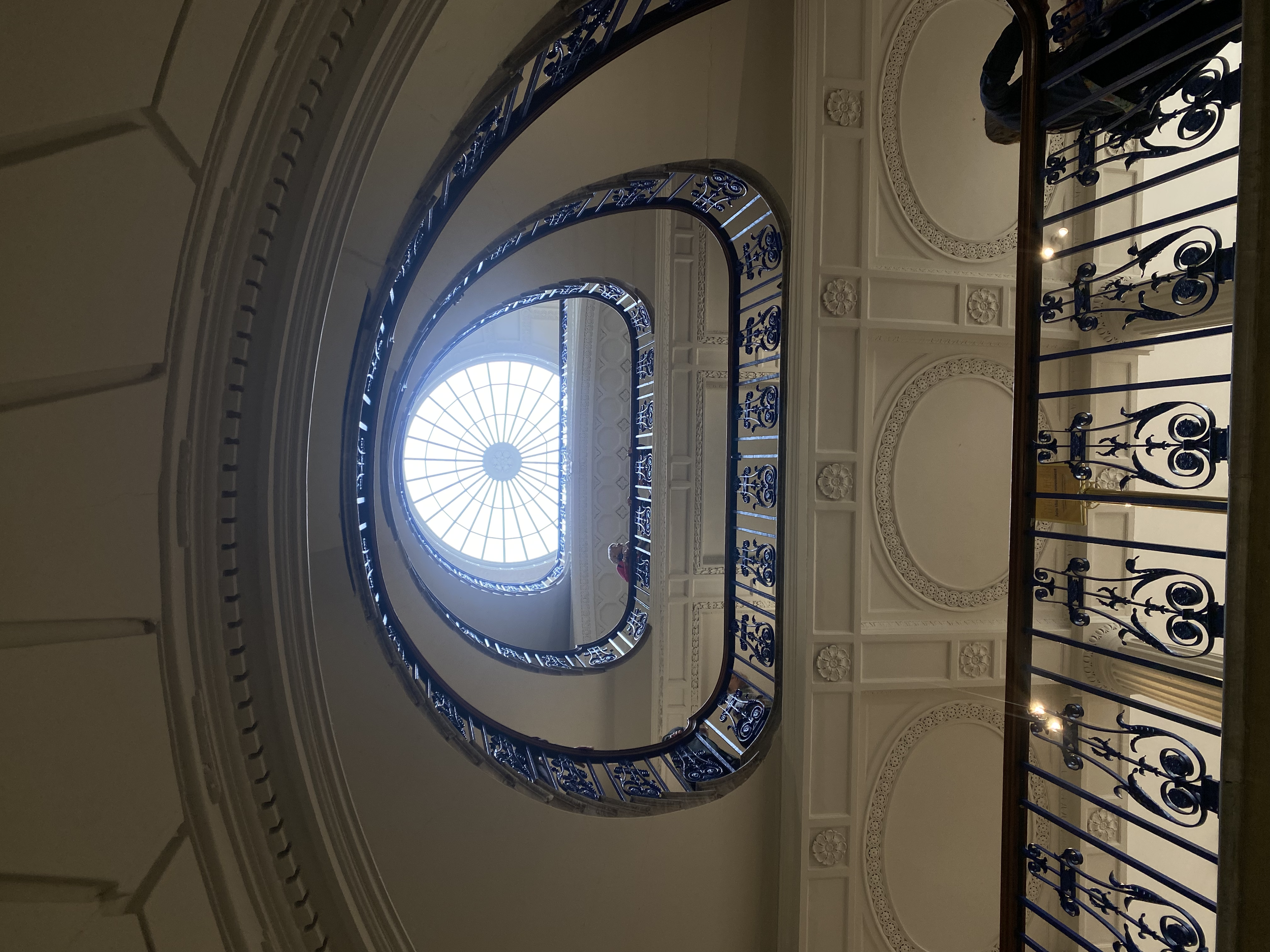 Looking up a spiral staircase with an ornate metal railing and a large circular skylight at the top.