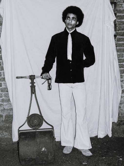 A man with an afro hairstyle wearing a dark shirt, light pants, and a scarf, standing next to a vintage grass cutting machine against a white sheet background.