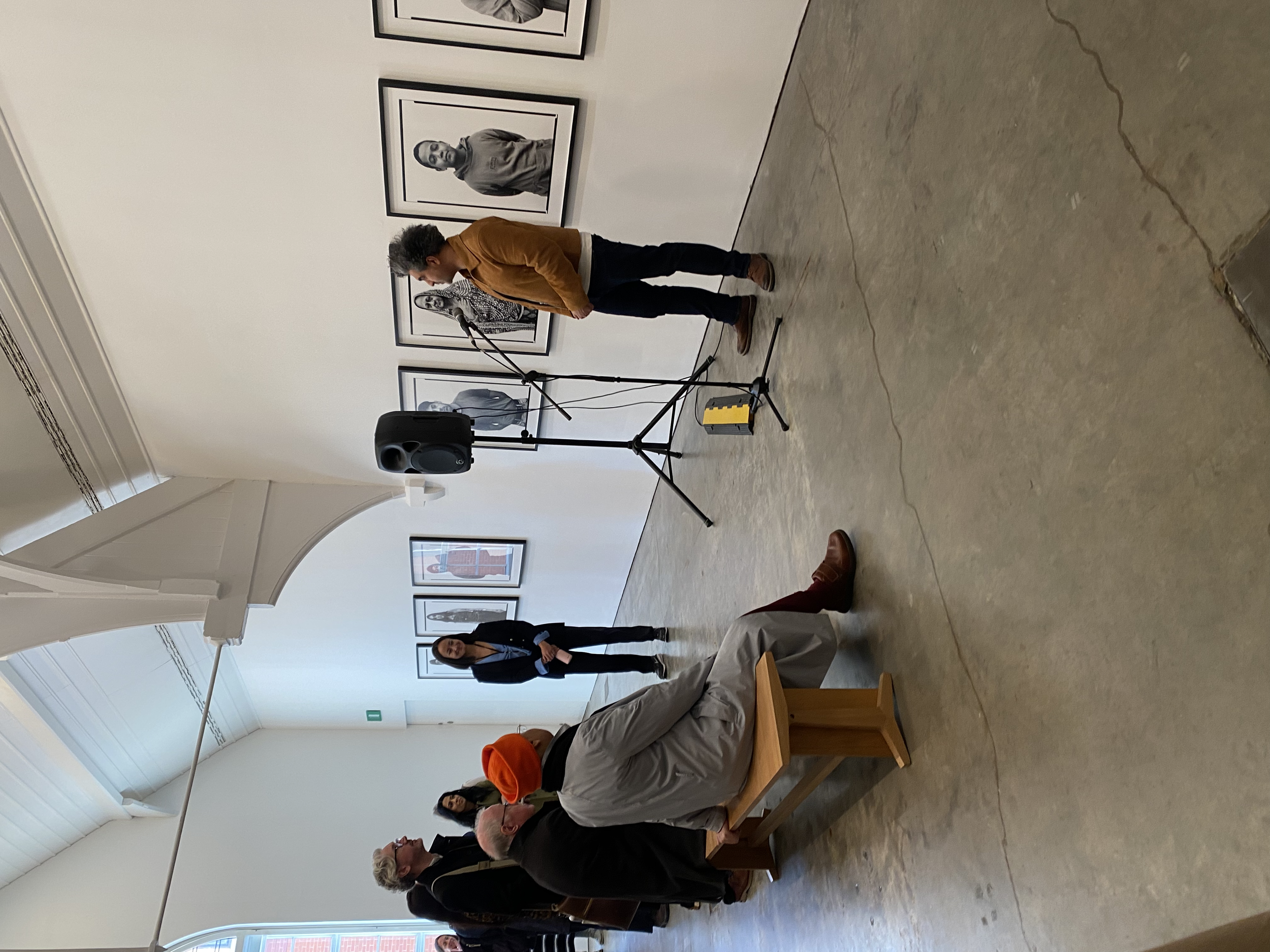 A gallery setting with a person speaking into a microphone, standing next to framed photographs on the wall. Several people are seated on benches facing the speaker, and a speaker system is visible on a stand. The room has a high ceiling and large wi