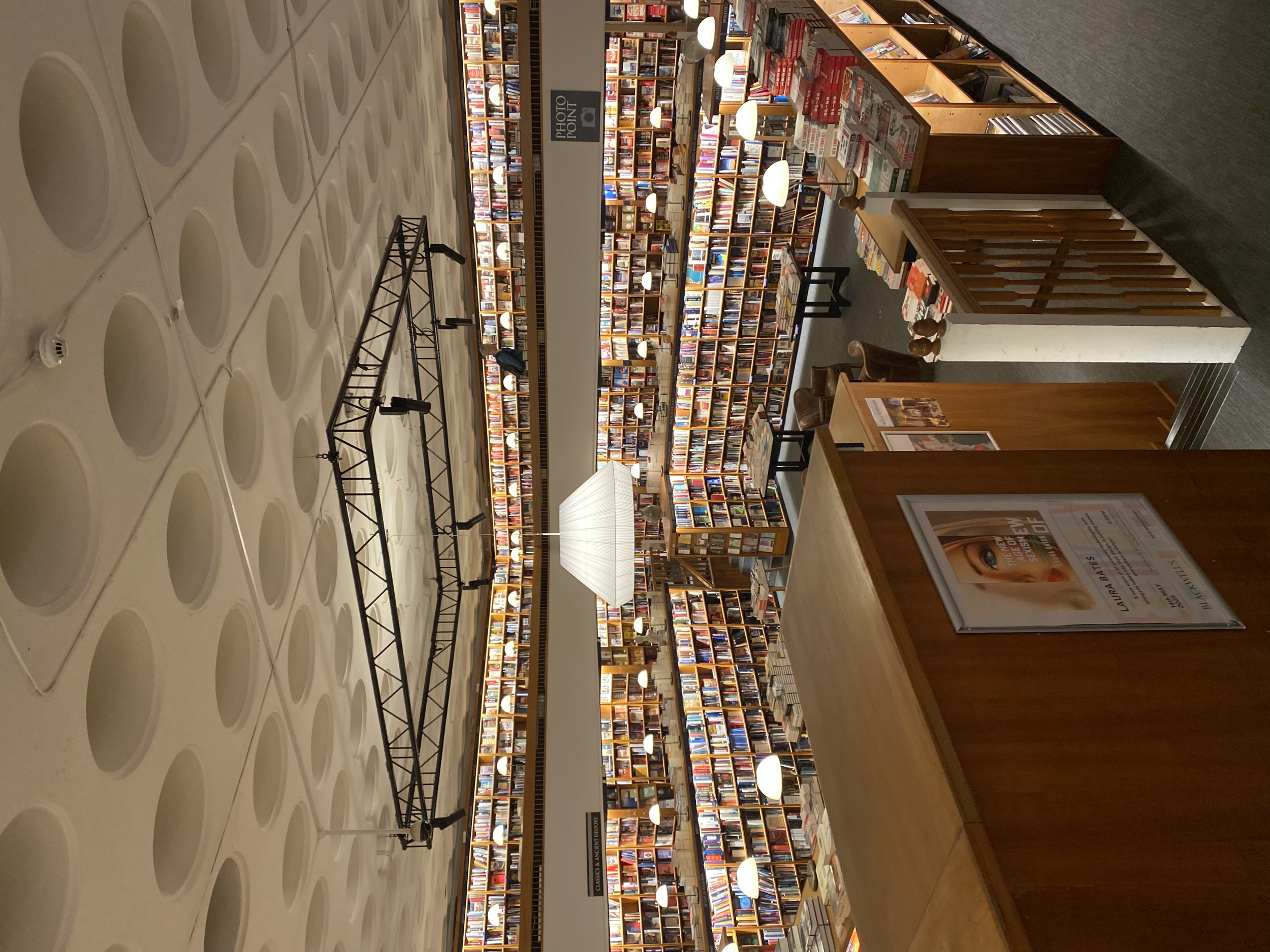 Interior of a large bookstore with bookshelves filled with books, a sign labeled "Photo Font," and wooden furniture. The ceiling has circular patterns, and there are hanging lights and tables for browsing.