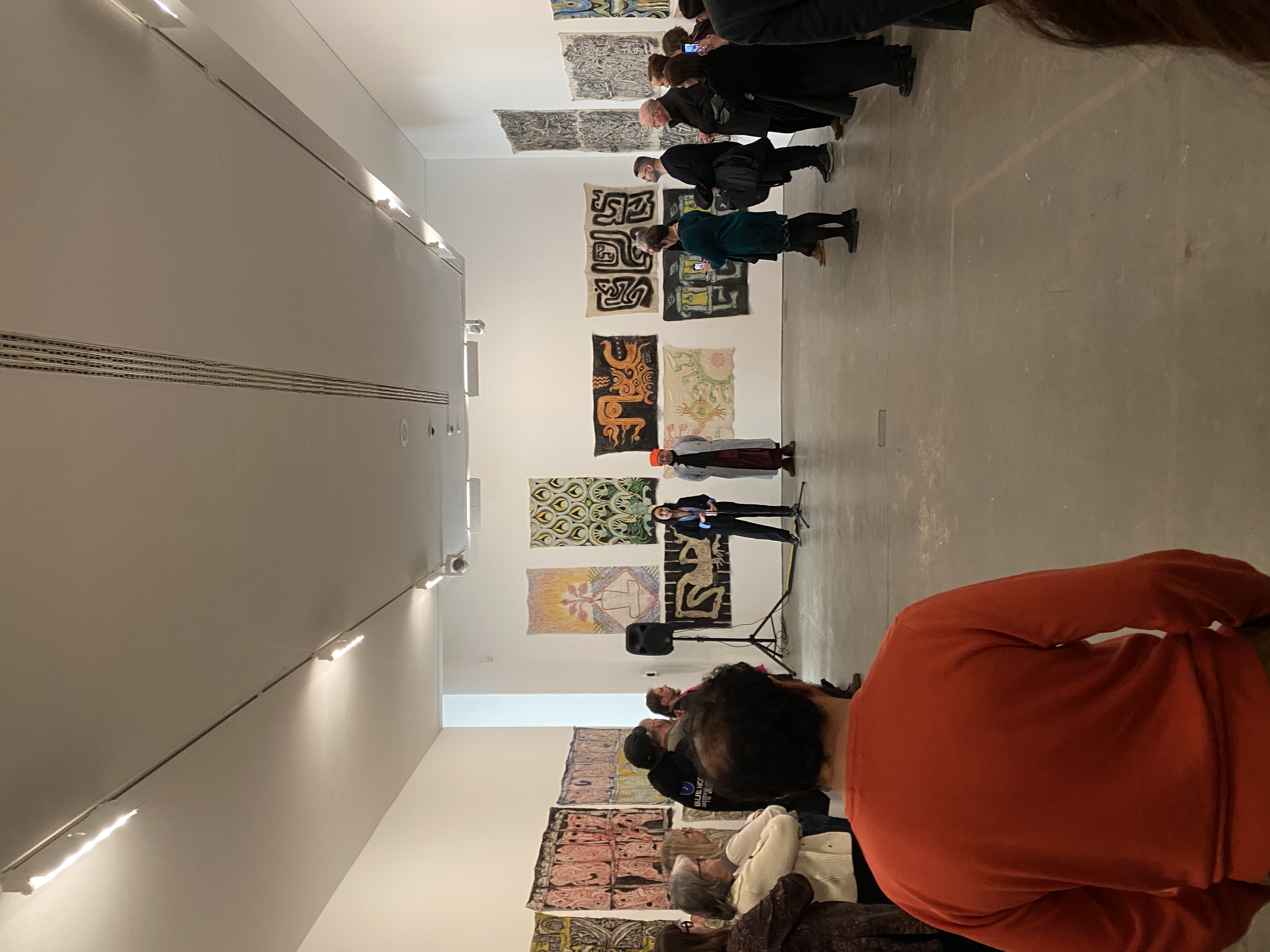 A gallery with people gathered, artwork displayed on the walls, and a person speaking at the front.