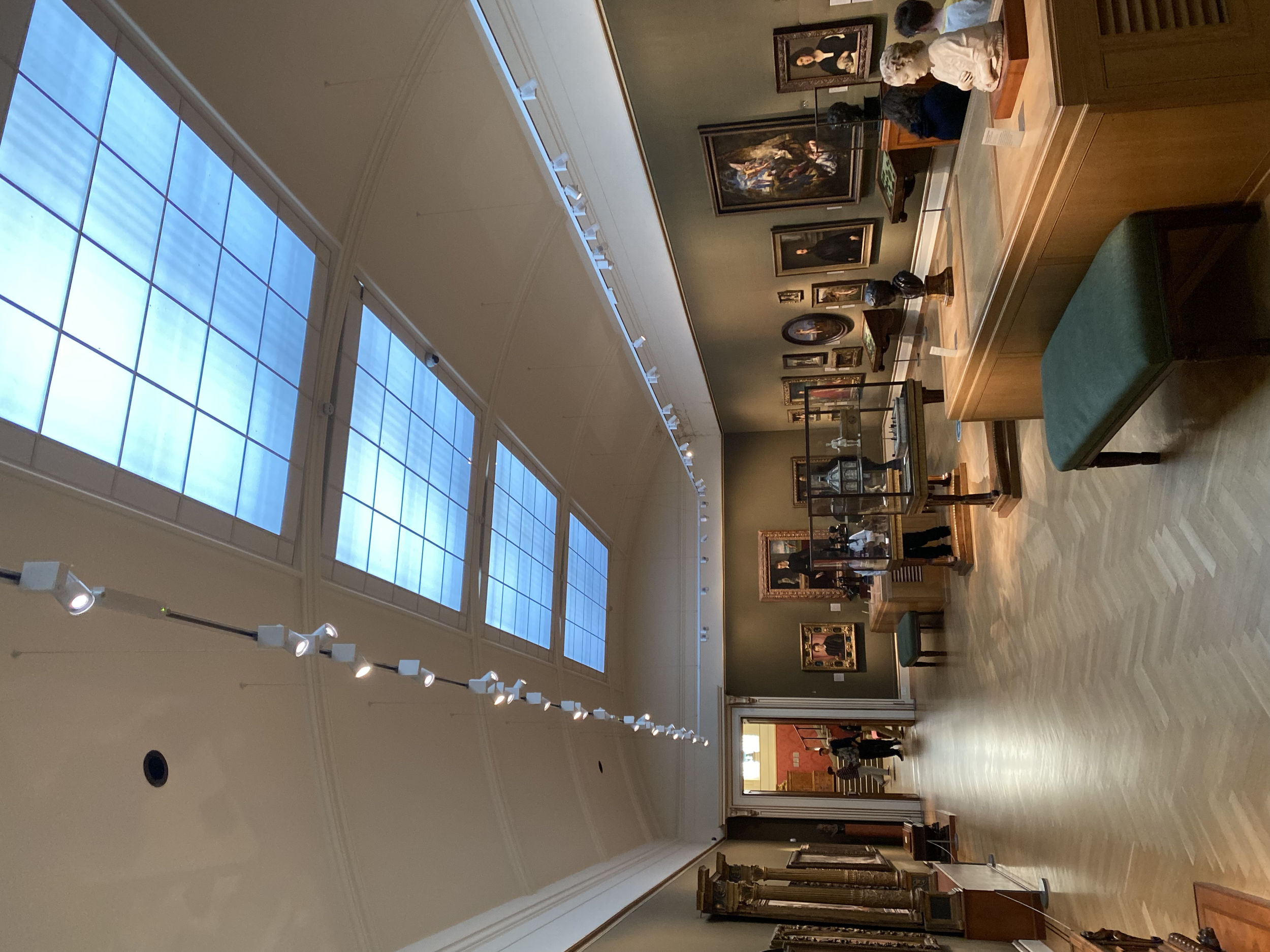 Art gallery interior with framed paintings, sculptures, and display cases under tall windows.