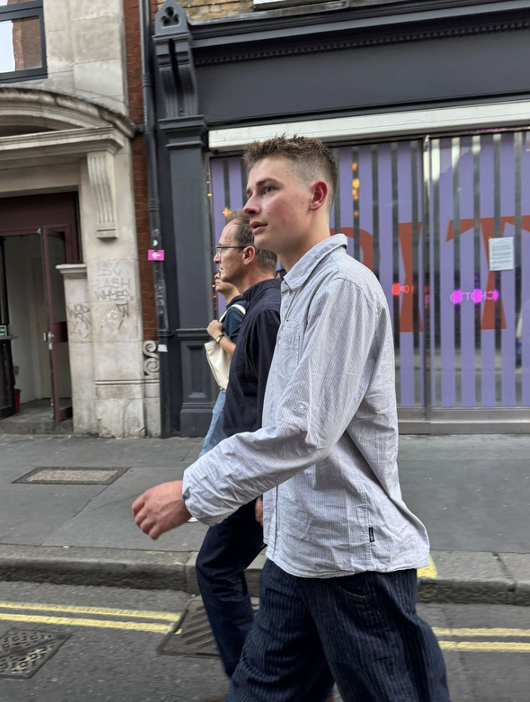 Three men walking on a city sidewalk, with the young man in the foreground wearing a light-colored shirt.